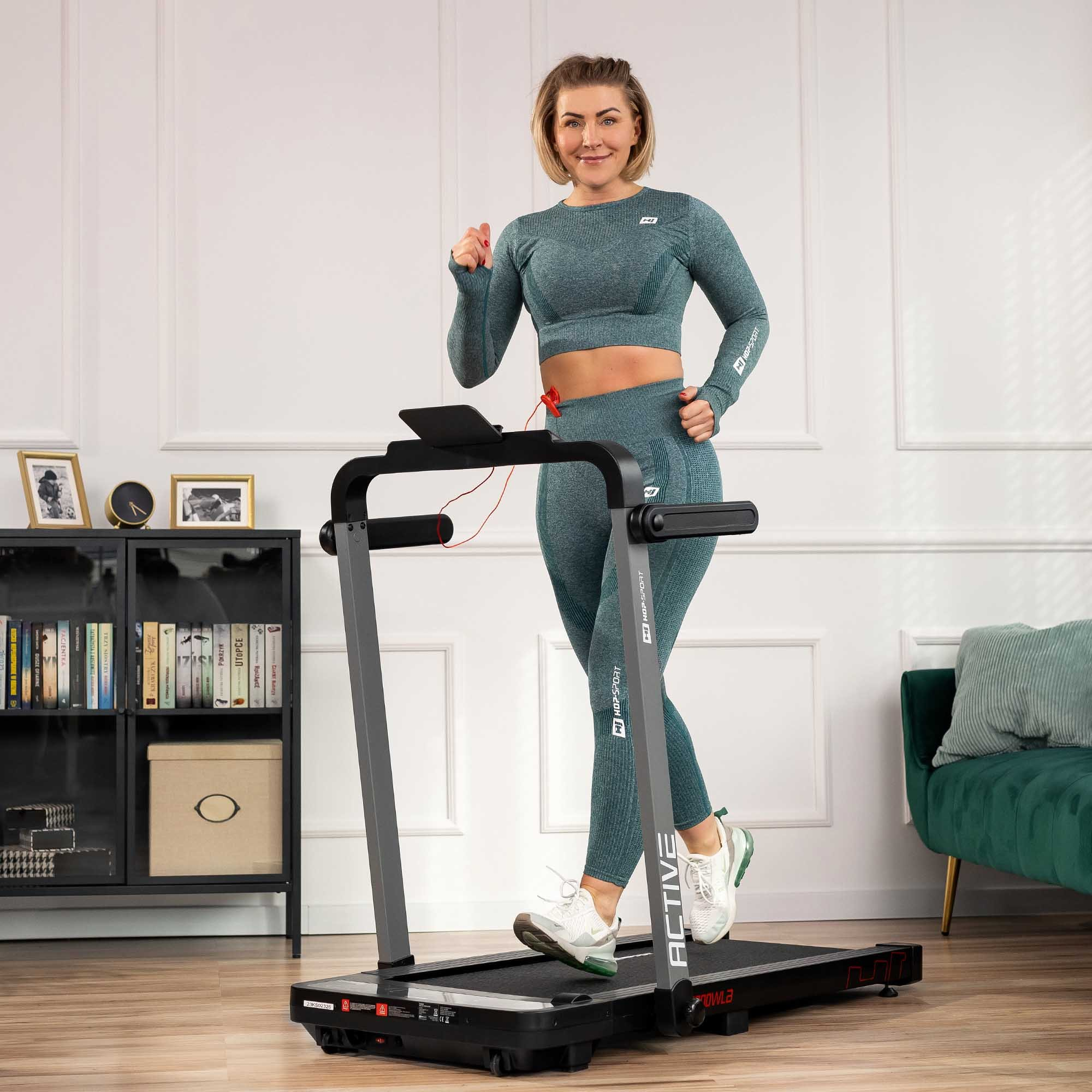 Eine Frau in grüner Sportbekleidung geht auf einem Laufband in einem hellen Raum mit Bücherregal und Sofa.