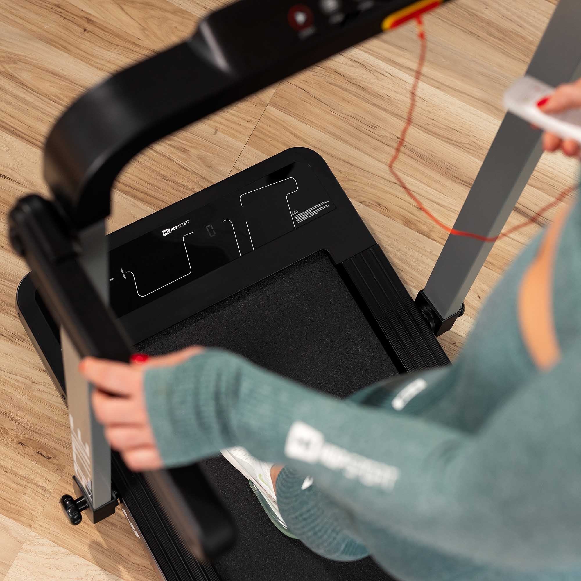 Eine Frau in grüner Sportbekleidung steht auf einem schwarzen Laufband auf einem Holzboden.