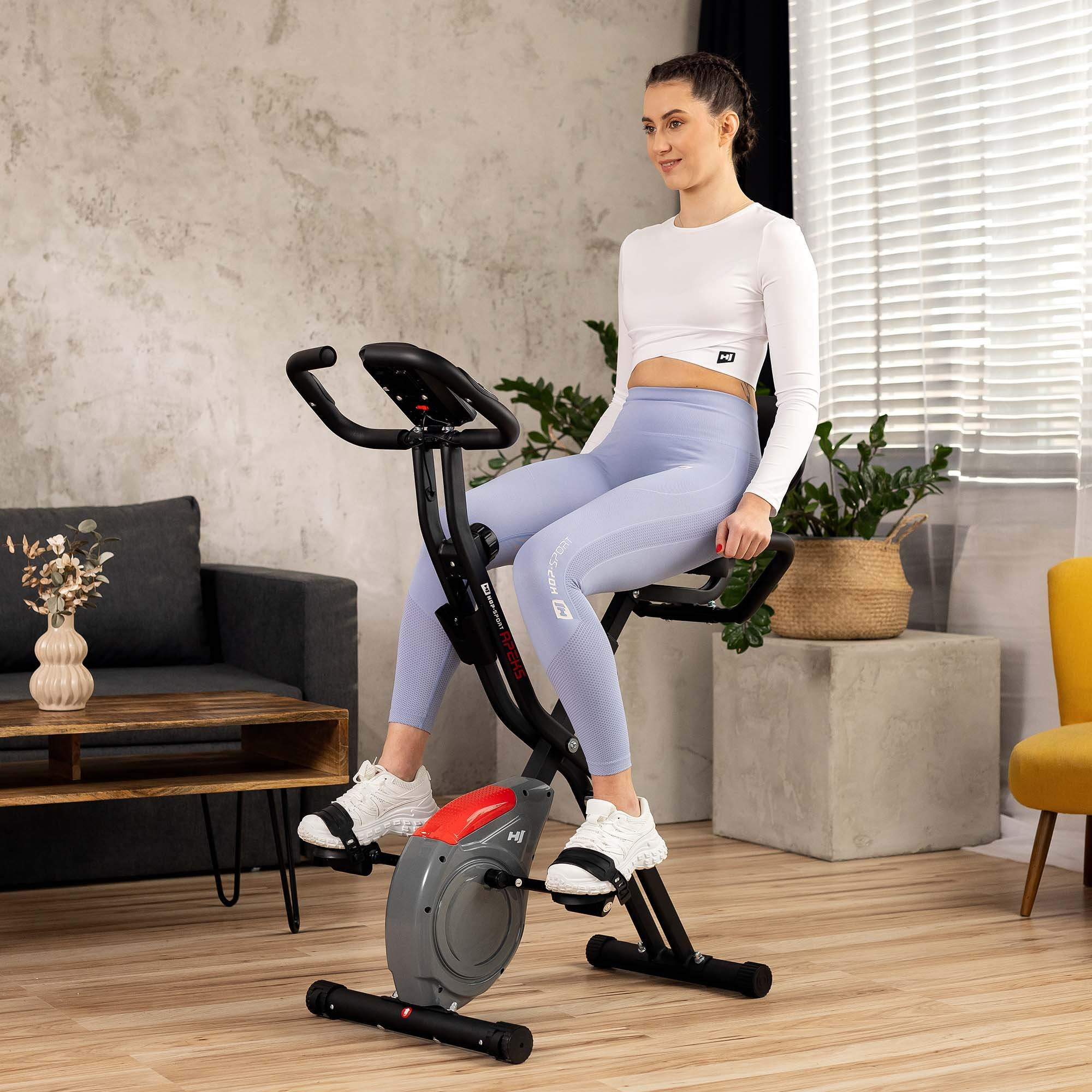 Eine Frau in weißem Oberteil und blauen Leggings trainiert in einem hellen Wohnzimmer auf einem Hometrainer.