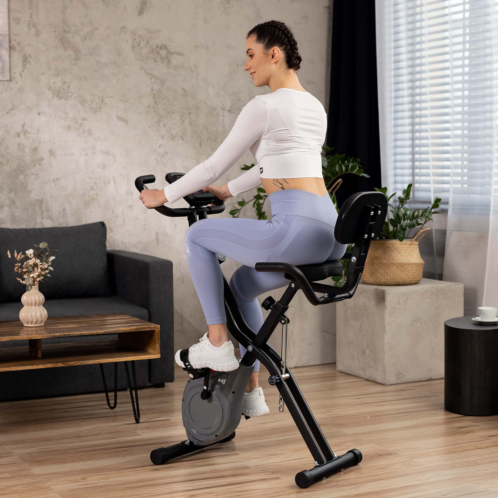 Eine Frau in weiß-blauer Sportkleidung trainiert auf einem Heimtrainer in einem Wohnzimmer.