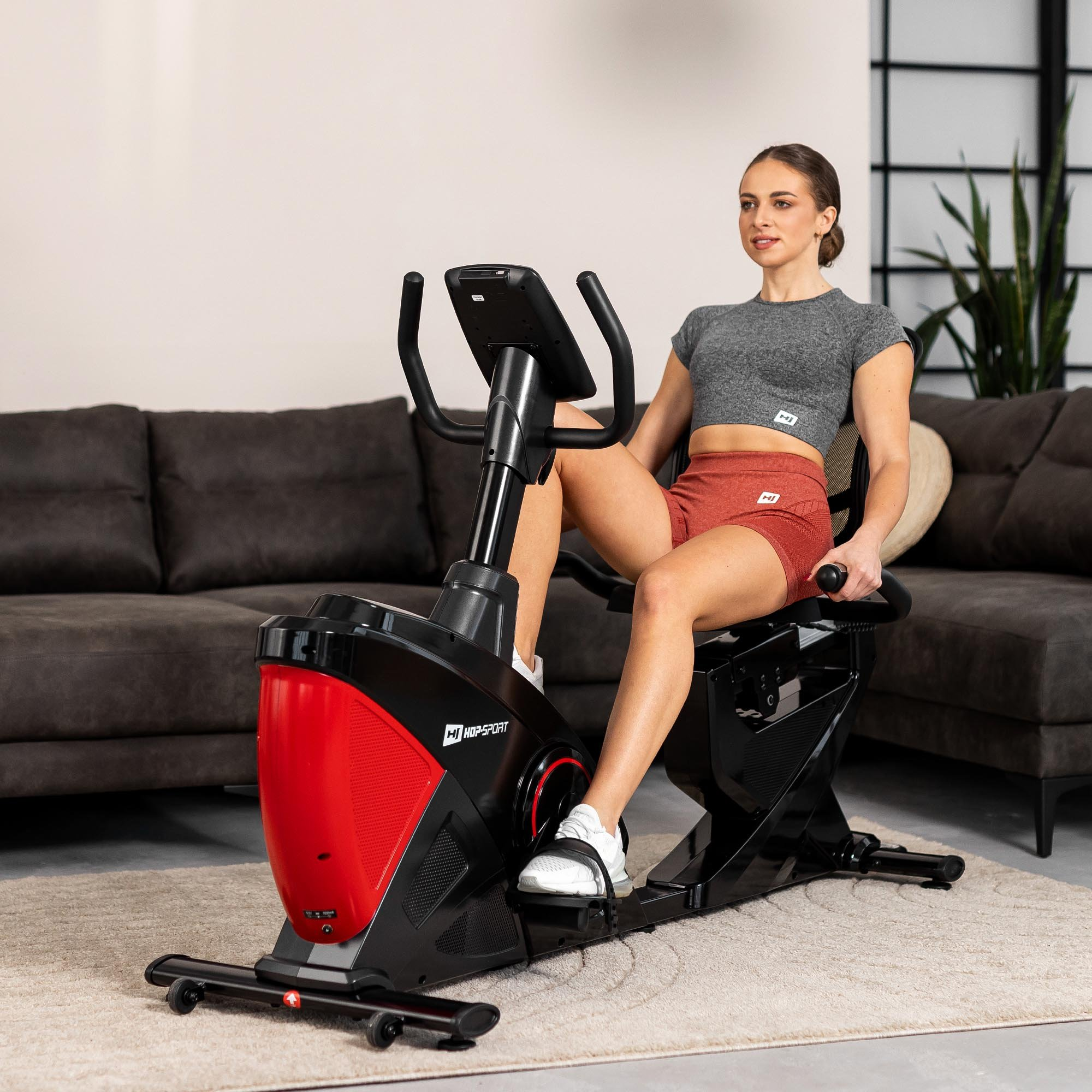 Eine Frau in grauem Crop-Top und roten Shorts trainiert auf einem schwarz-roten Liegeheimtrainer im Wohnzimmer.