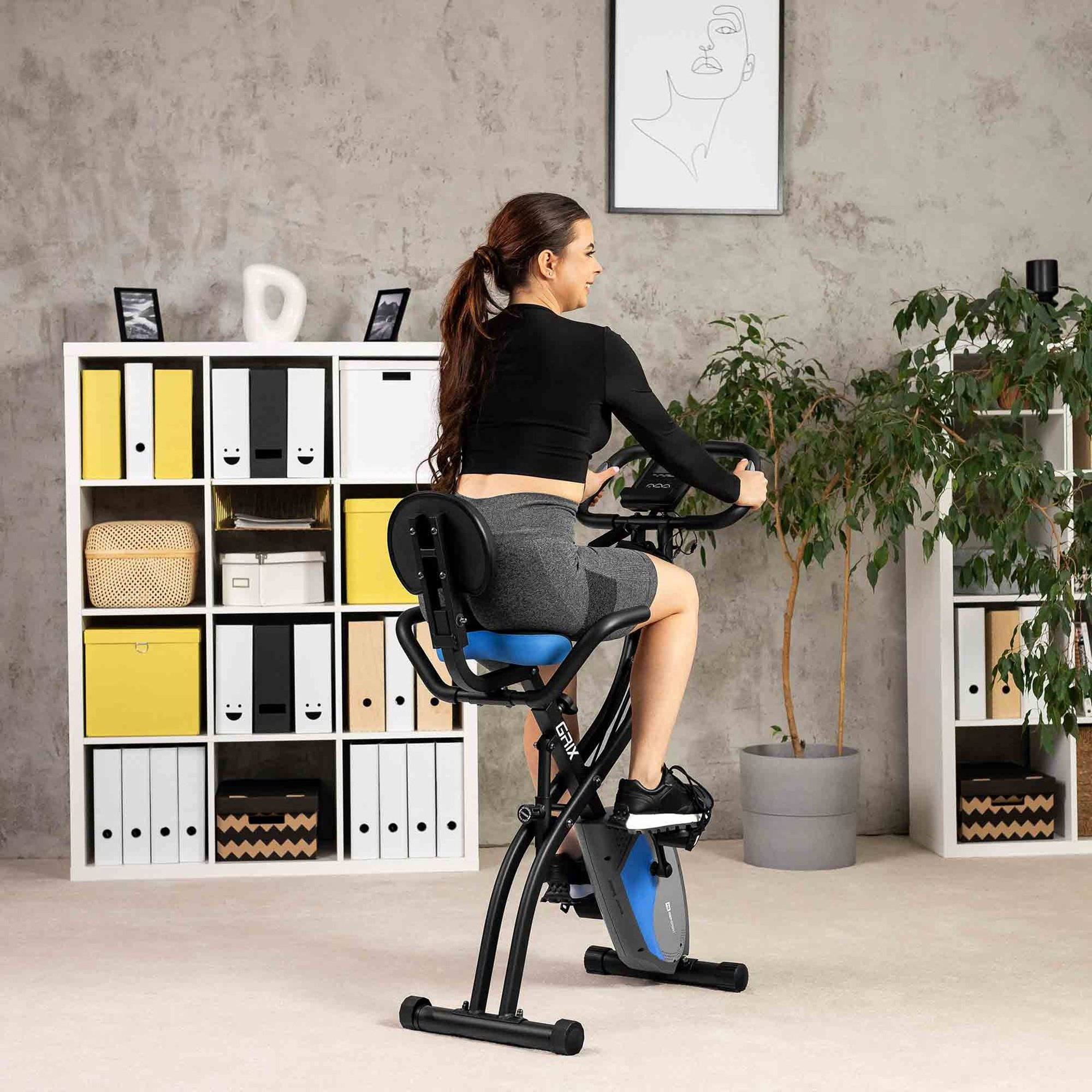 Eine Frau trainiert in einem hellen Raum auf einem schwarzen und blauen Heimtrainer, umgeben von Regalen und Pflanzen.