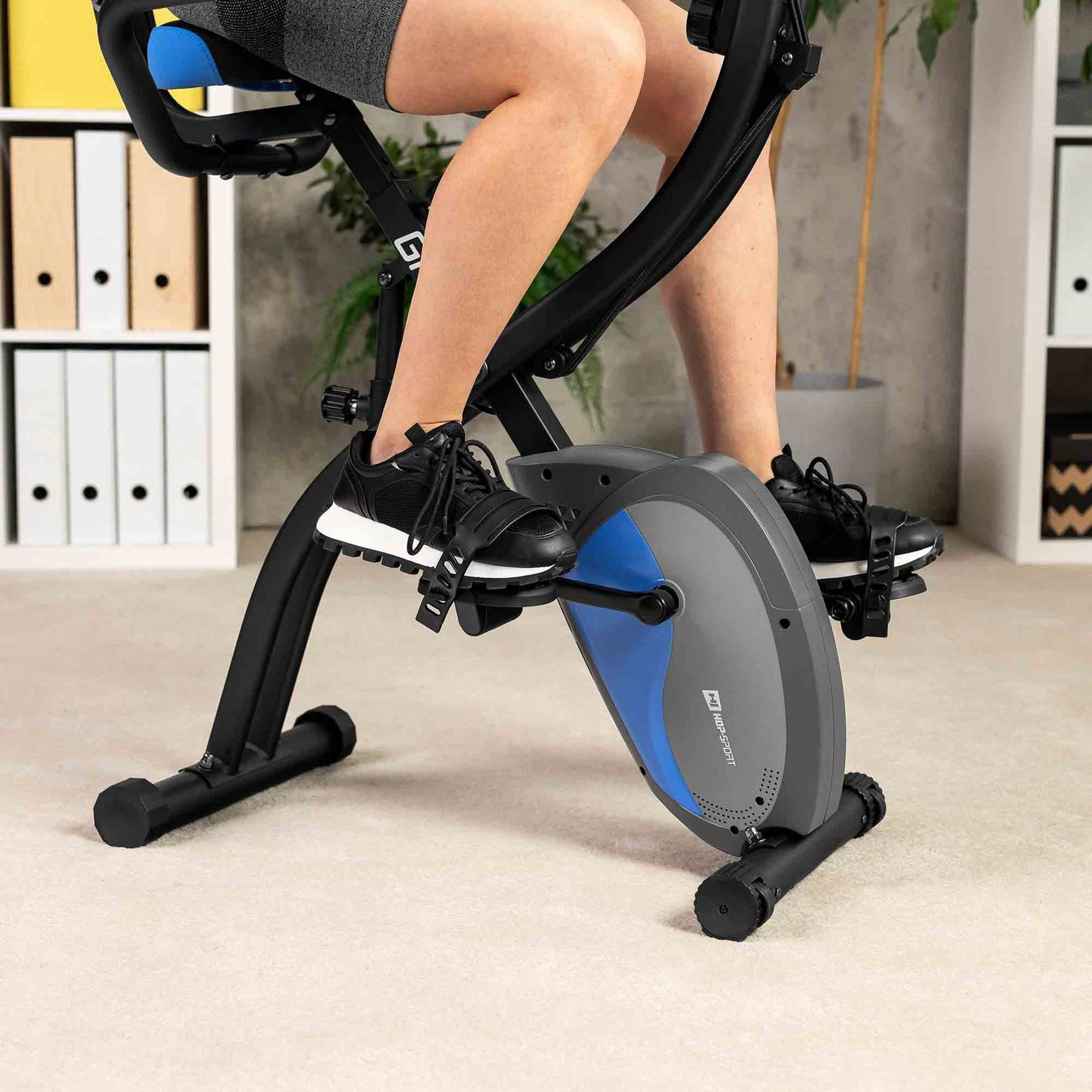 Eine Person sitzt auf einem schwarzen Heimtrainer mit blauem Akzent in einem Raum mit Regalen.