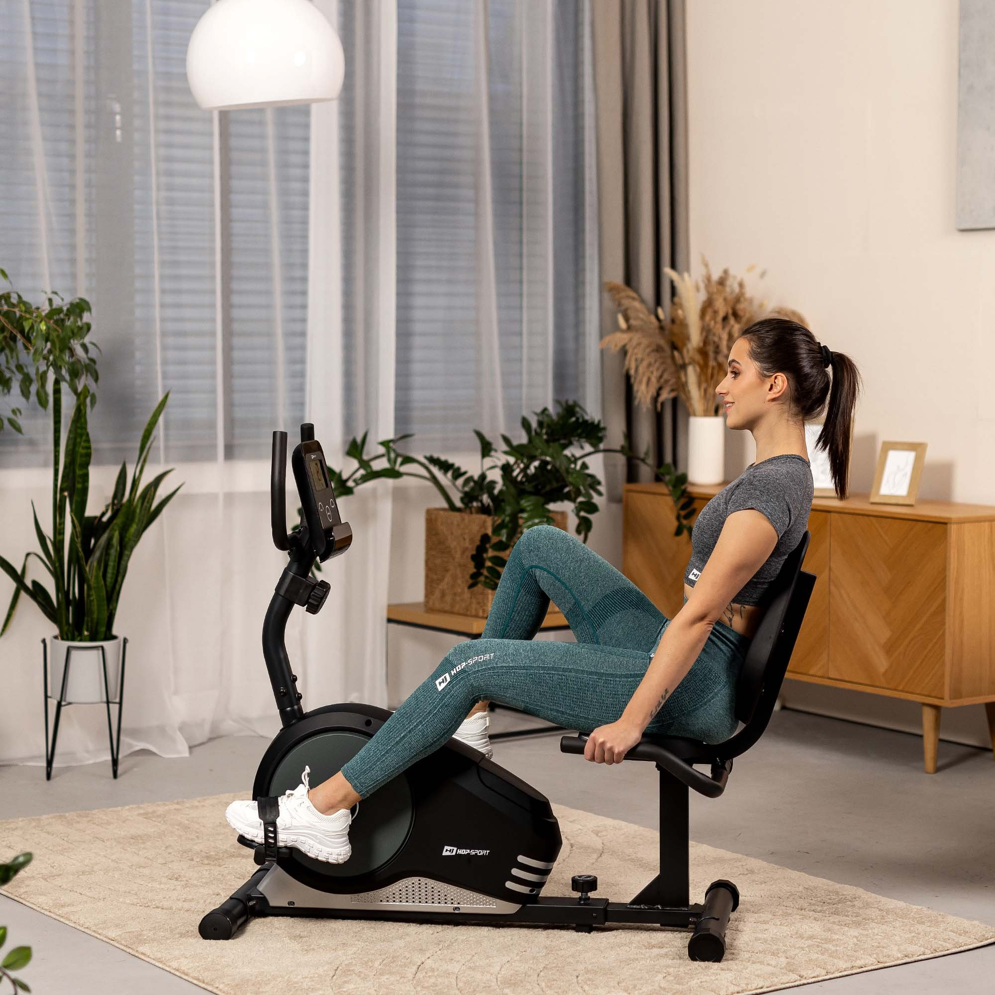 Eine Frau in Sportkleidung trainiert zu Hause auf einem schwarzen Liegeheimtrainer.