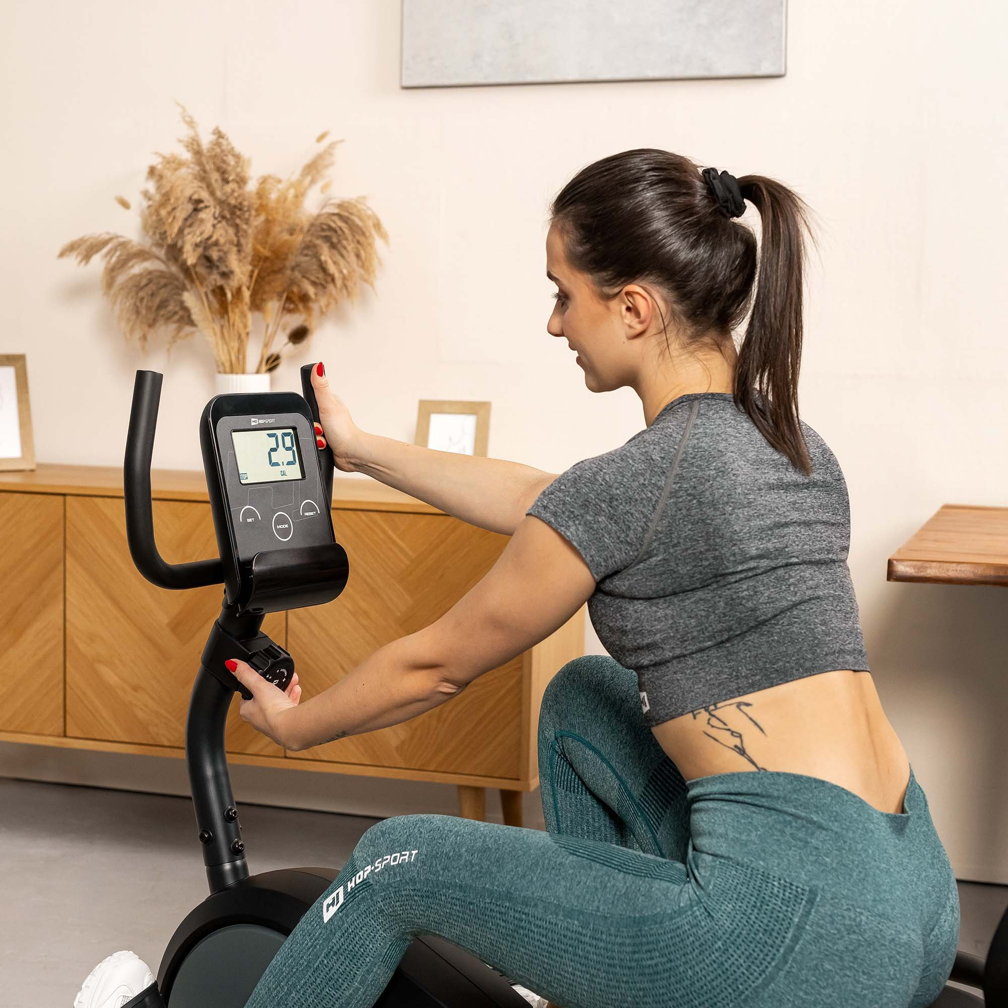 Eine Frau in grauer Sportkleidung trainiert auf einem Hometrainer in einem Raum mit Holzzimmerschrank und Pflanzen.
