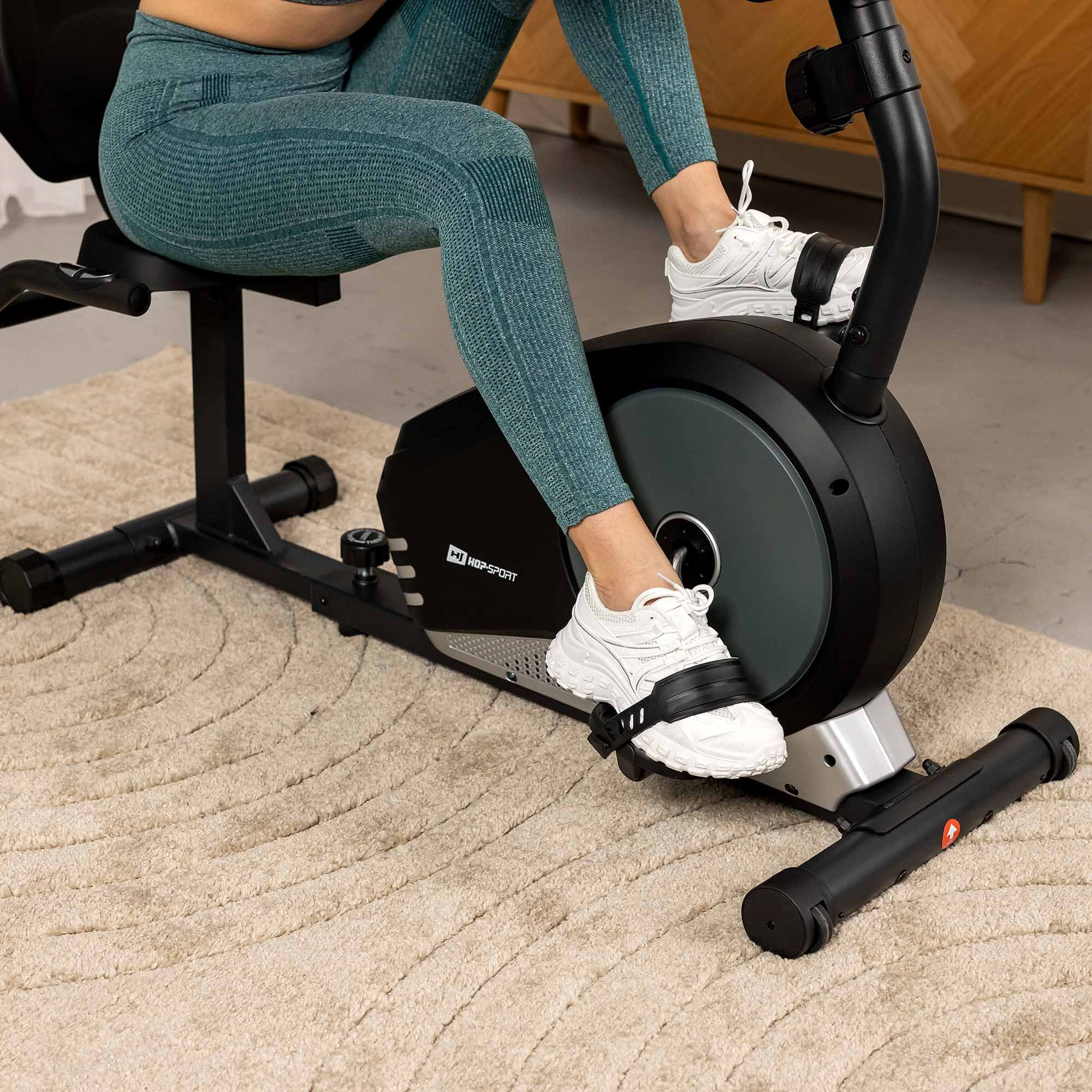 Person in blauen Leggings radelt auf einem schwarzen Heimtrainer auf einem beigen Teppich.