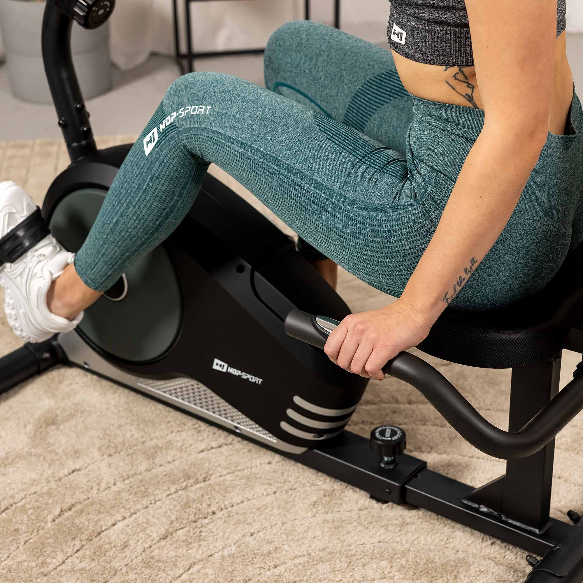 Eine Frau in grüner Sportkleidung sitzt auf einem schwarzen Heimtrainer und trainiert drinnen.