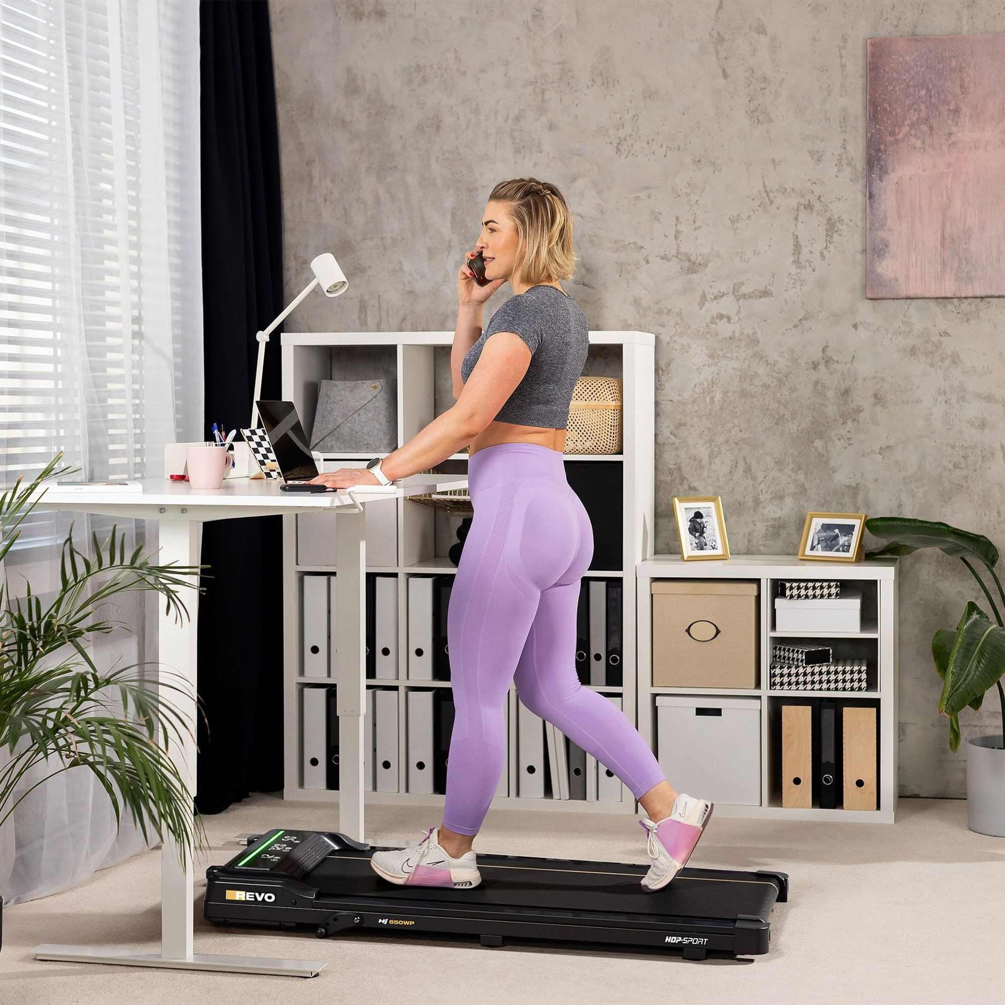 Eine Frau in grauem Top und lila Leggings geht auf einem Laufband und telefoniert an einem Stehpult.