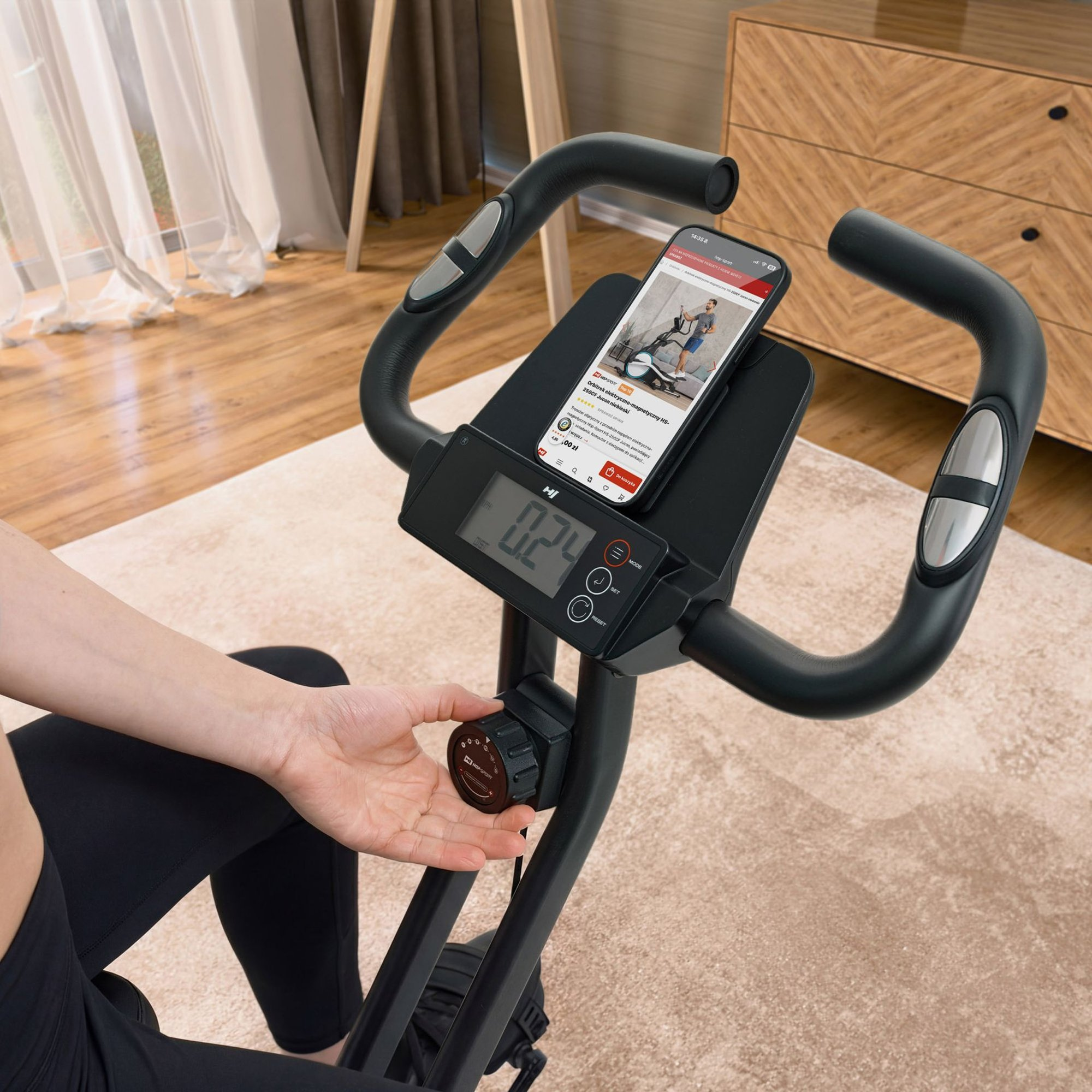 Eine Person stellt den Widerstandsknopf an einem Heimtrainer mit Digitalmonitor und Smartphonehalterung ein.