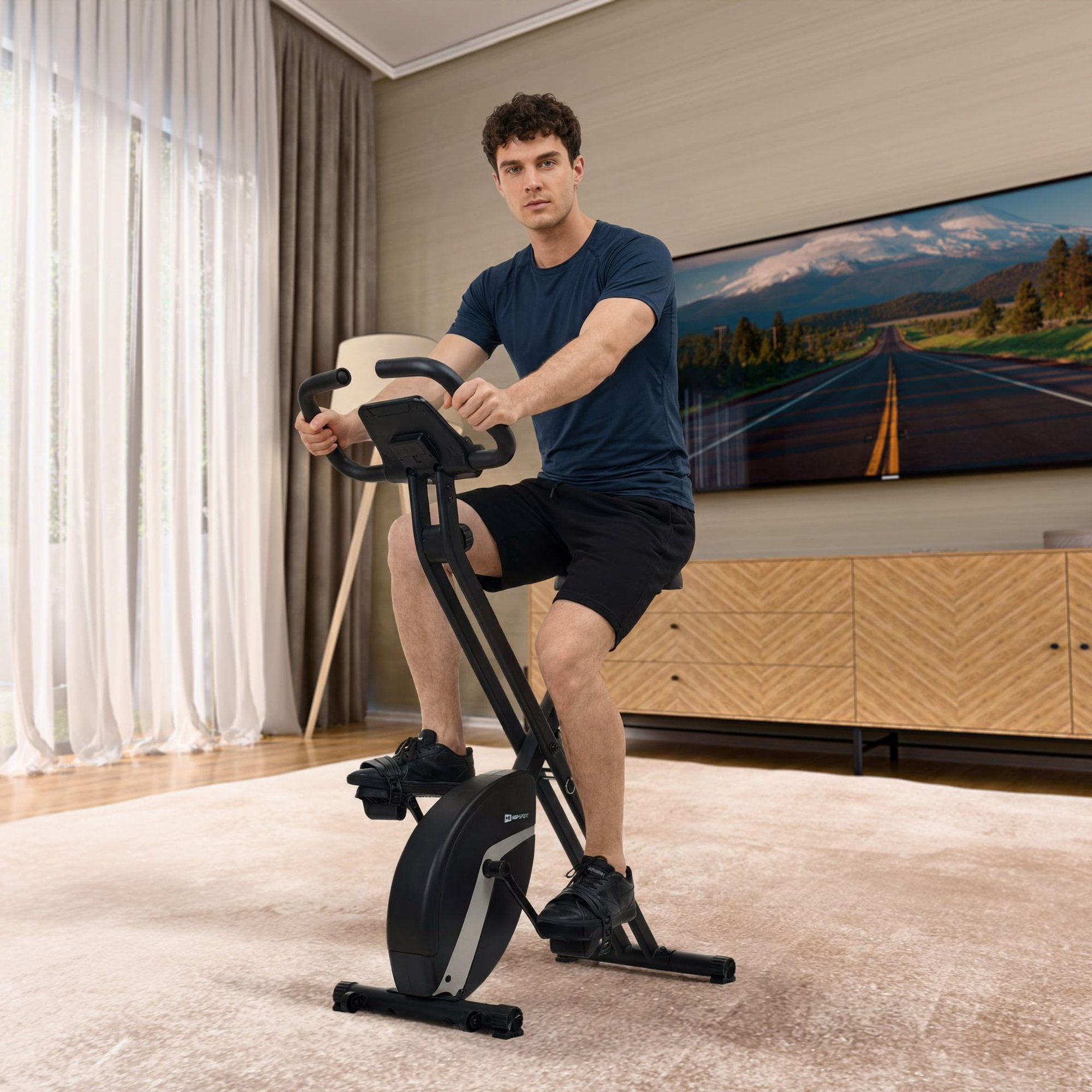 Ein Mann sitzt in einem Wohnzimmer auf einem schwarzen Heimtrainer und blickt in die Kamera.