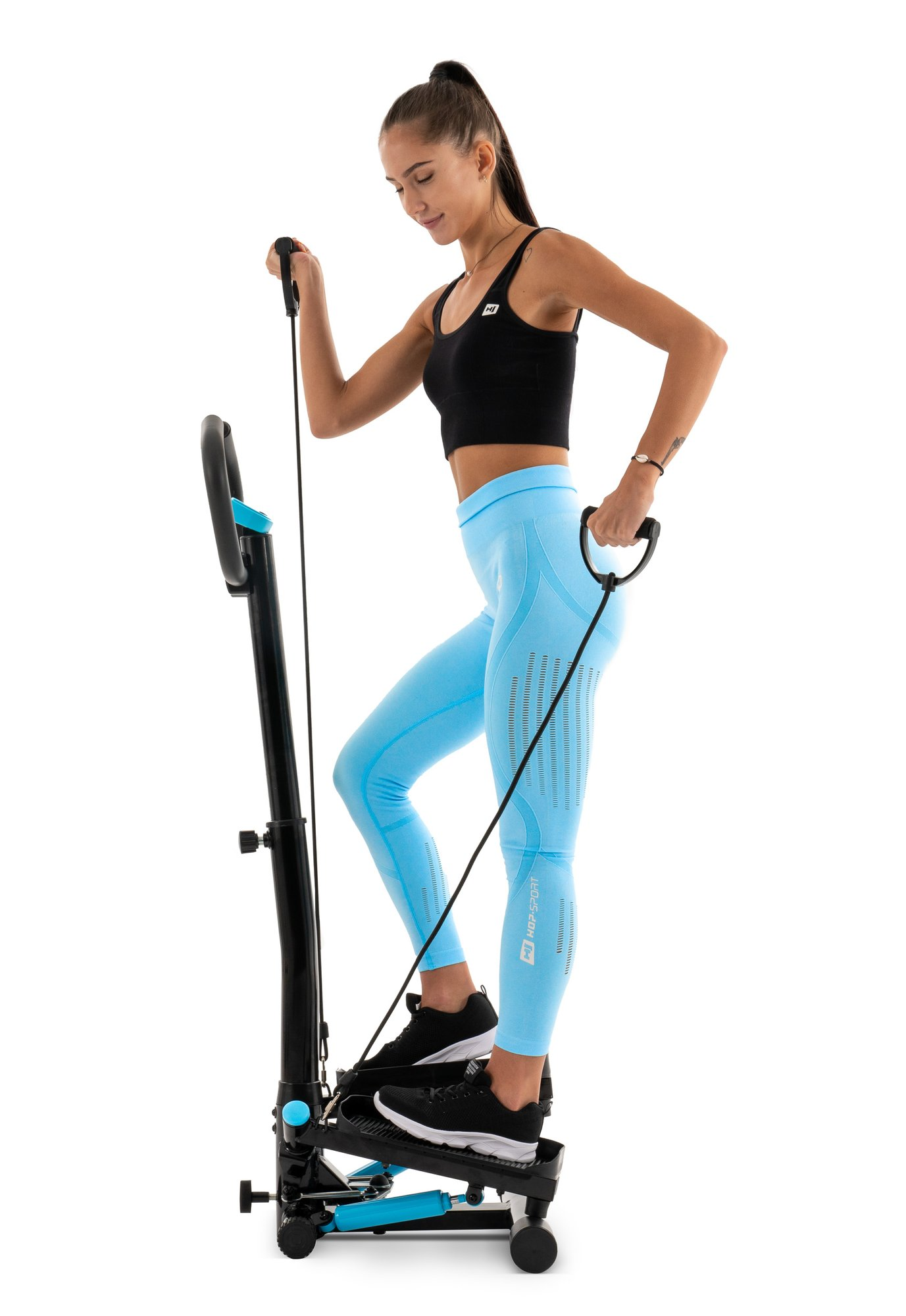Eine Frau im schwarzen Oberteil und blauen Leggings trainiert auf einem schwarzen Stepper mit Bändern vor weißem Hintergrund.