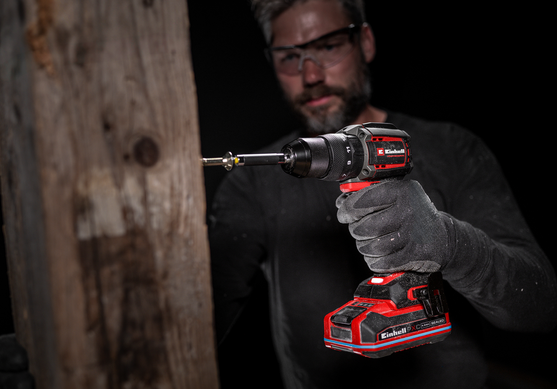 Un artigiano con occhiali di protezione usa un trapano a batteria rosso per avvitare una trave di legno.