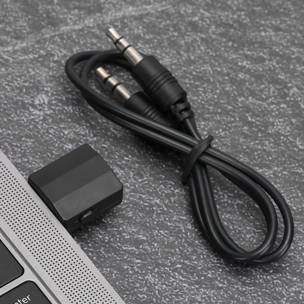 Ein schwarzes Audiokabel und ein USB-Empfänger liegen auf einer grauen Oberfläche neben einem Laptop.