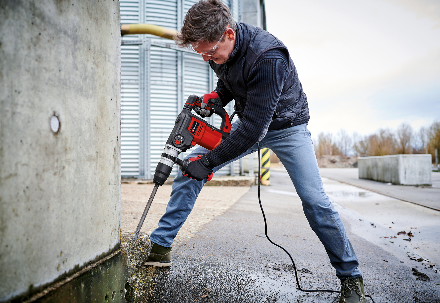 Ein Mann bohrt im Freien mit einem roten Bohrhammer in ein Betonfundament.
