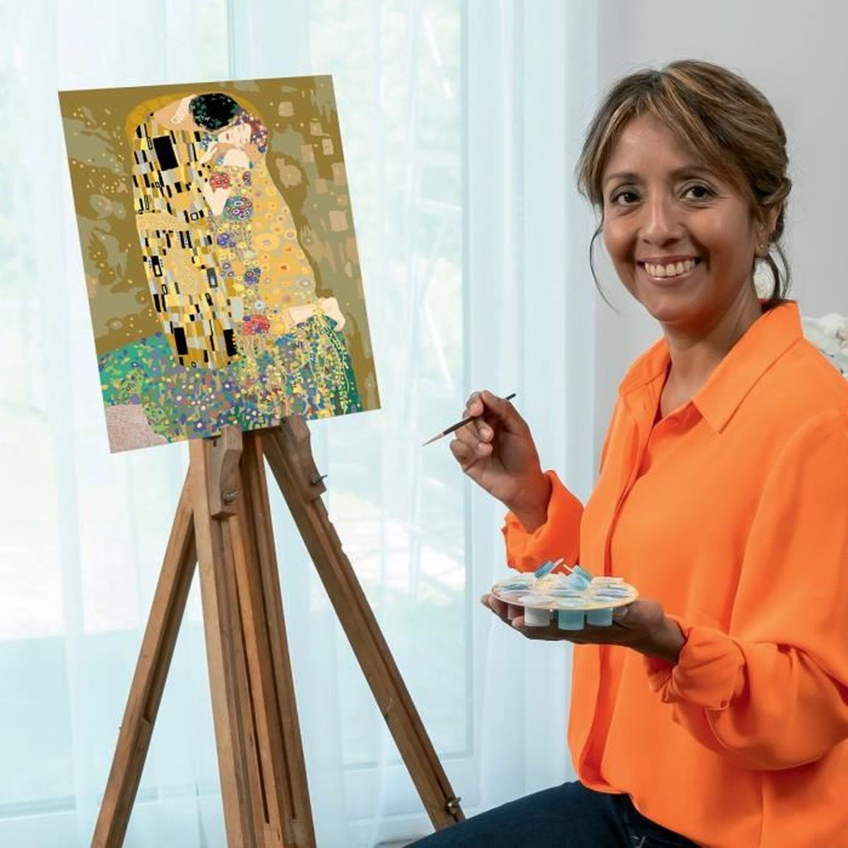 Eine lächelnde Frau in Orange malt vor einer Leinwand mit einem Bild im Stil von Klimts Der Kuss.