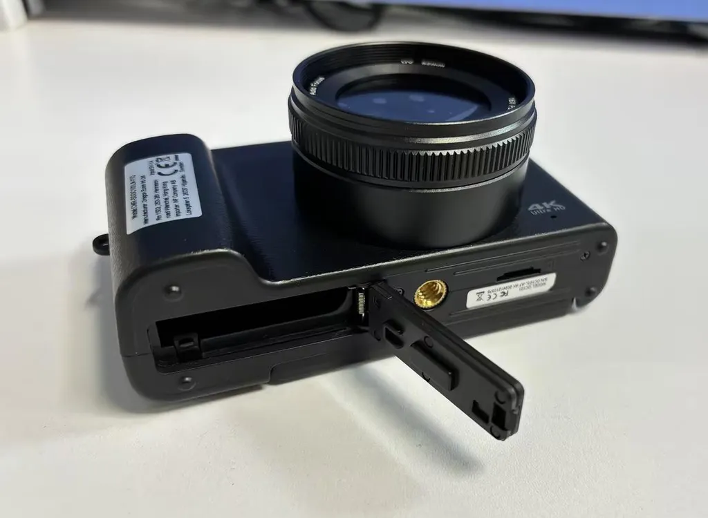 Eine schwarze Digitalkamera liegt auf einem weißen Tisch, die Batterieklappe ist geöffnet.
