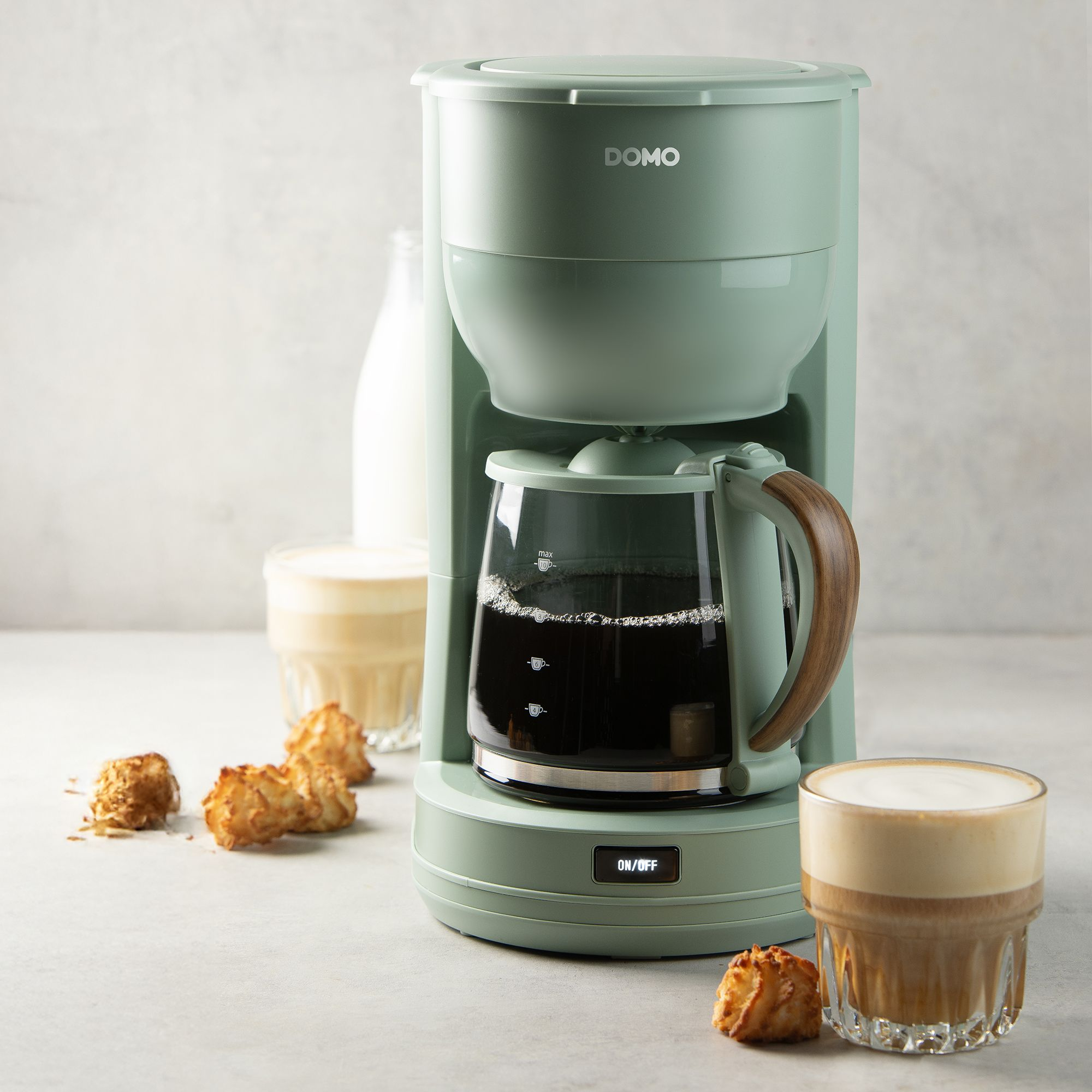 Grüne Filterkaffeemaschine mit Glasanae, Kaffeetasse und Gebäck auf grauem Tisch.