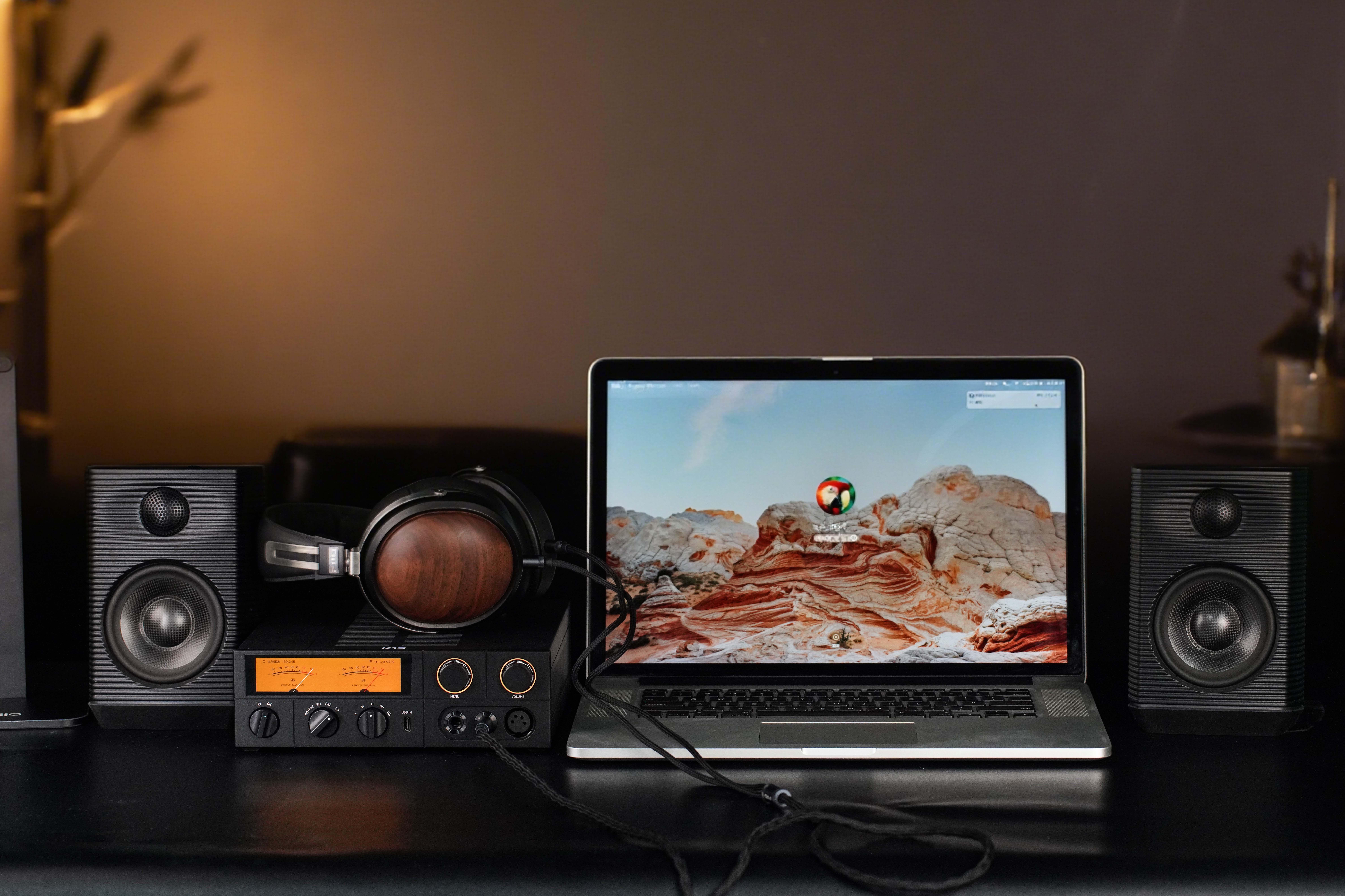 Un escritorio con un portátil, auriculares con orejeras de madera sobre un amplificador y dos altavoces negros.