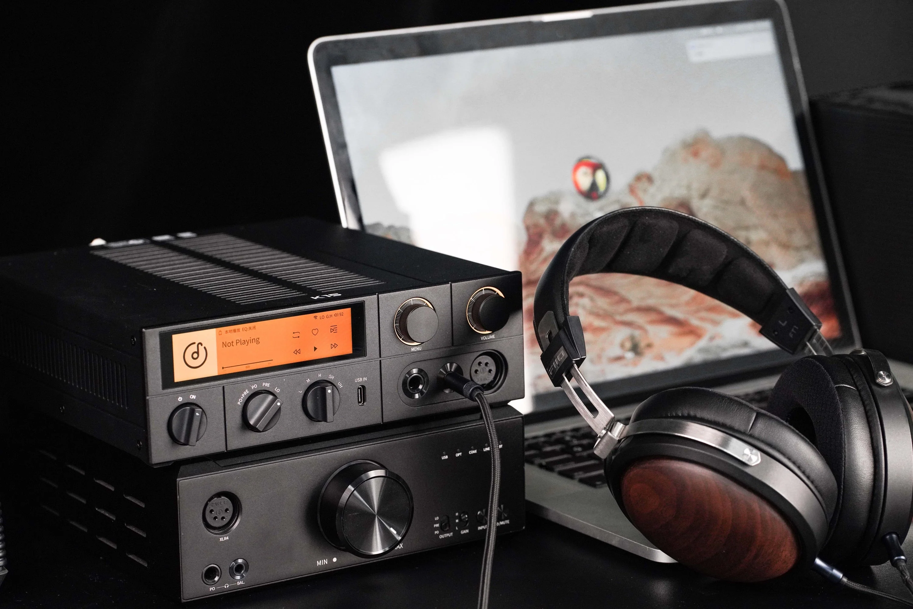 Amplificador de audio negro con pantalla, auriculares con detalles en madera y un portátil al fondo.