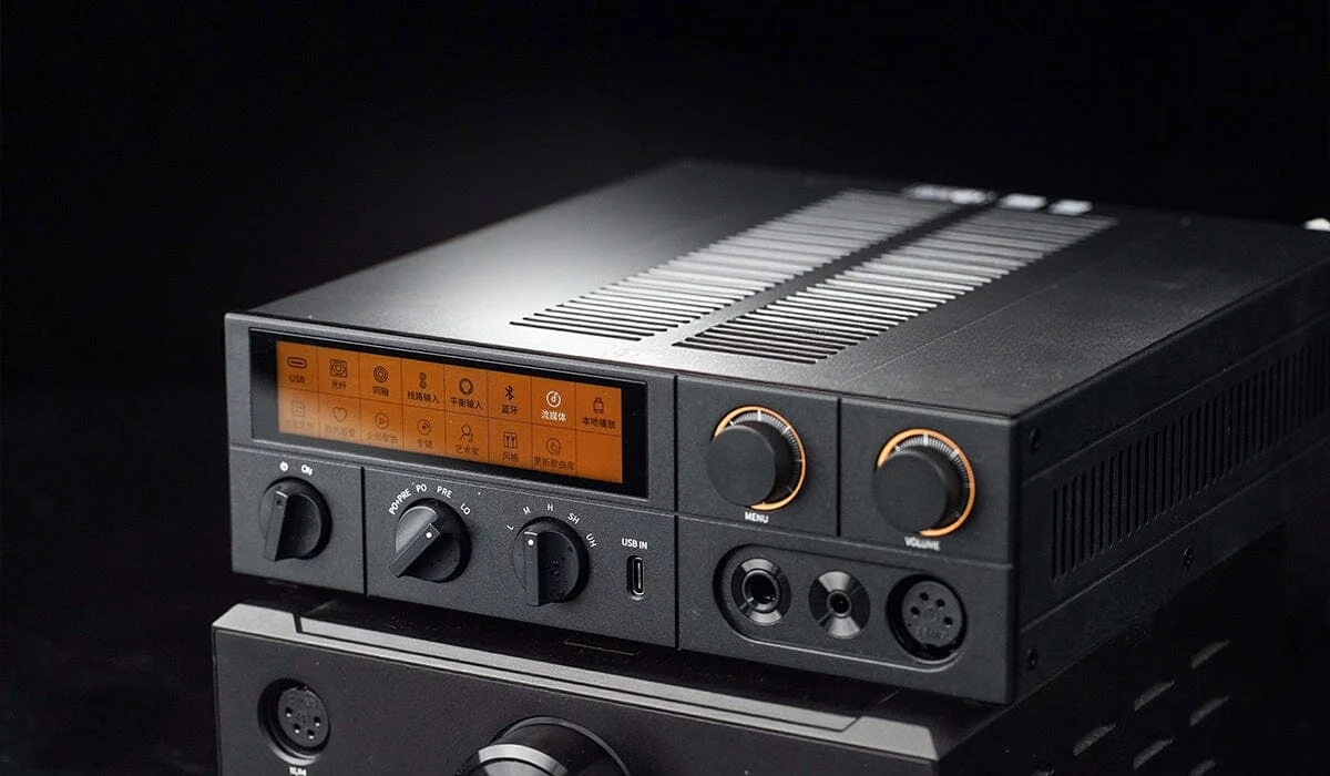 Amplificador de audio negro con pantalla naranja, perillas de control y puertos de audio sobre fondo oscuro.