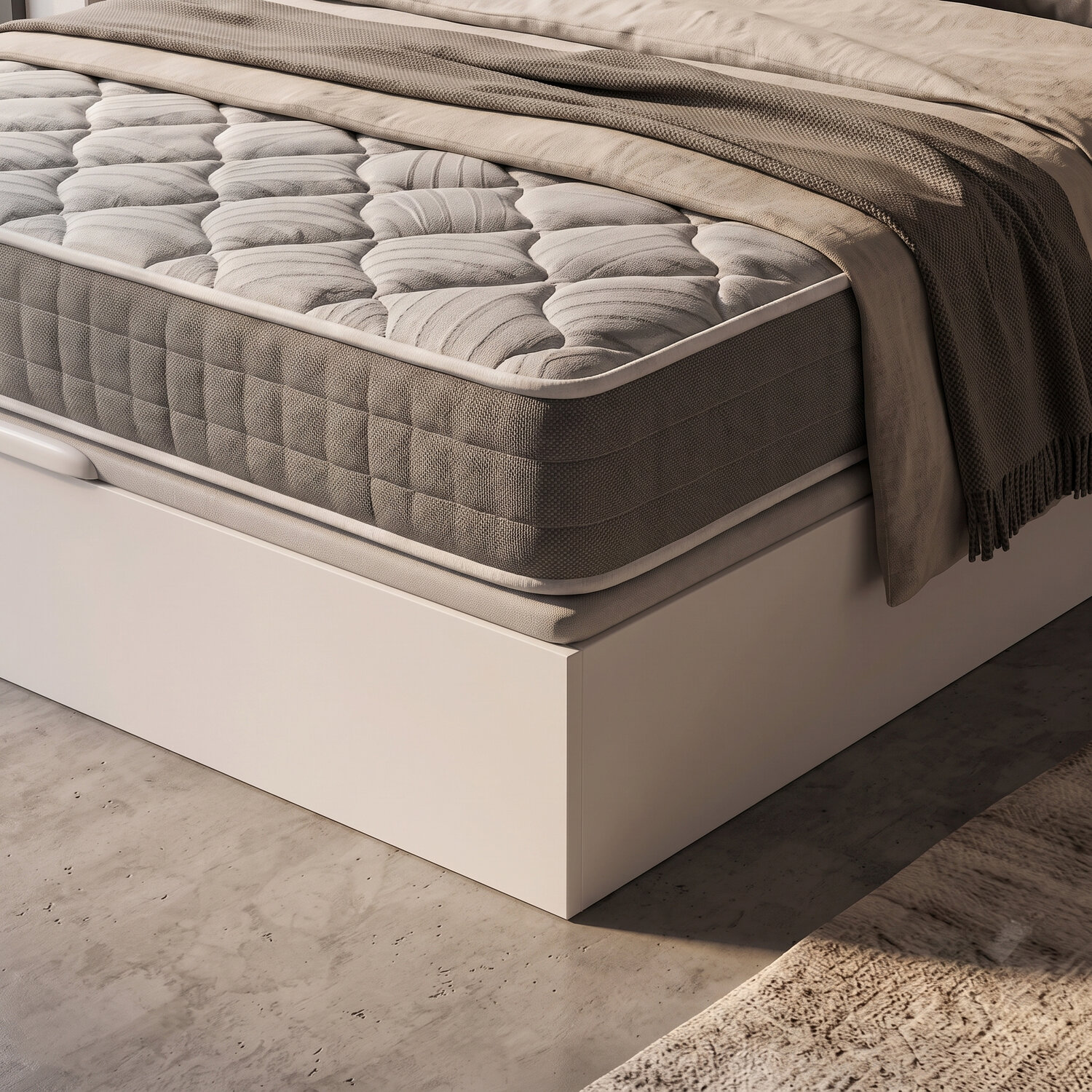 Primer plano de una cama con colchón acolchado, ropa de cama beis y estructura blanca en suelo de hormigón.