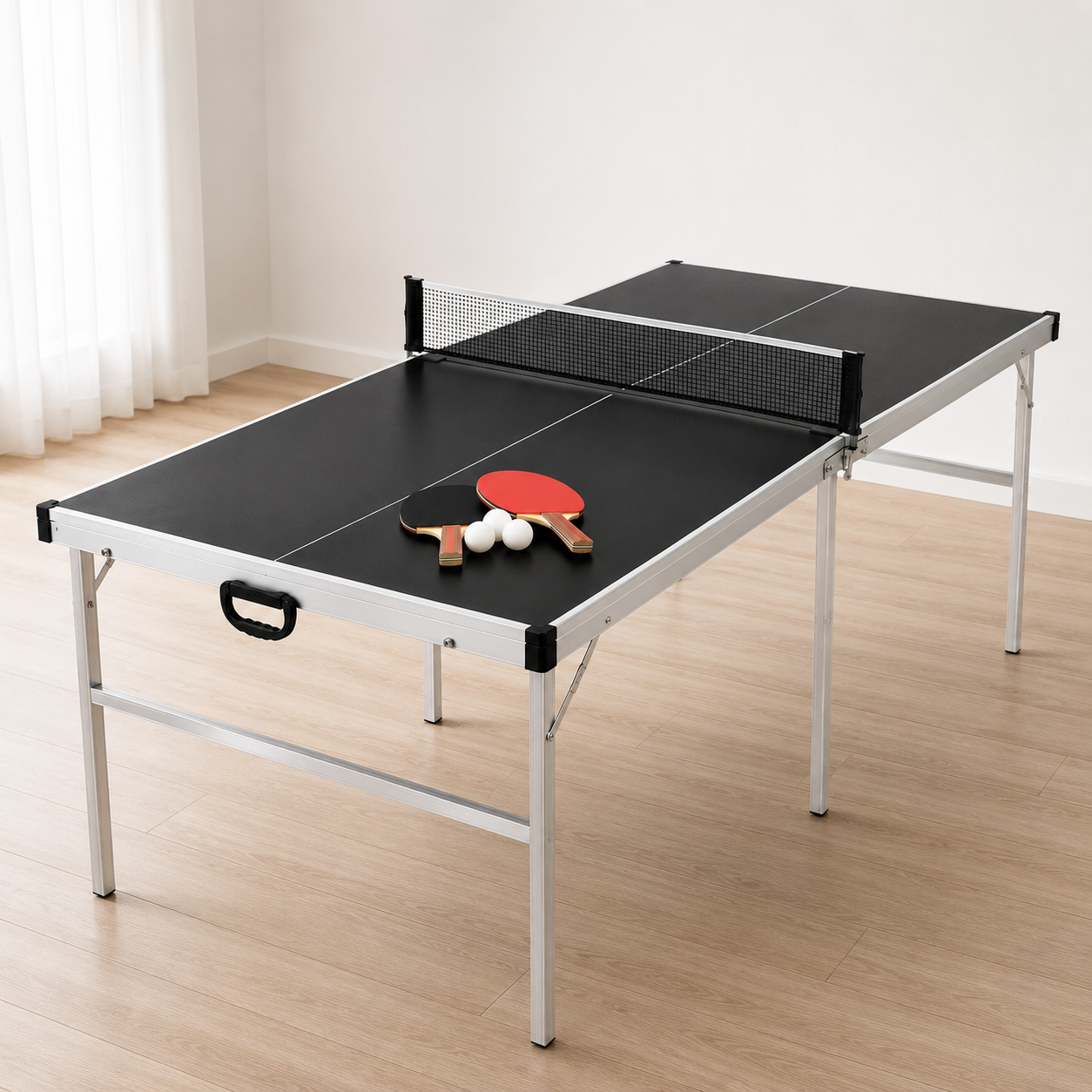 Una mesa de ping pong negra con red, dos paletas y tres pelotas sobre suelo de madera en una habitación luminosa.