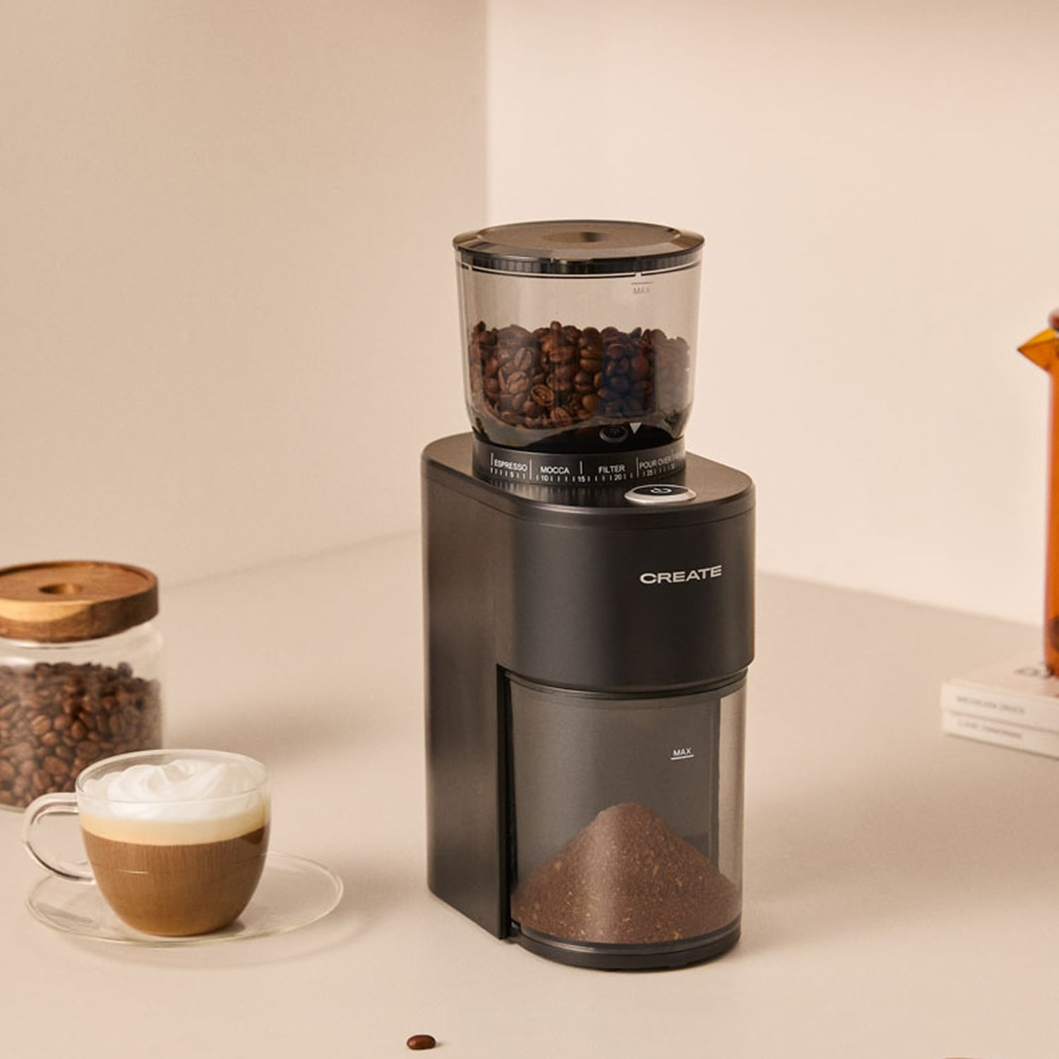 Un molinillo de café negro sobre una mesa con granos y café molido, al lado de una taza de capuchino y un frasco.