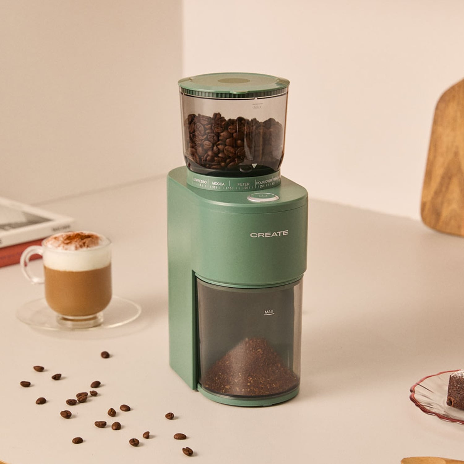 Molinillo de café verde en una mesa, lleno de granos y café molido. A la izquierda hay una taza de café con espuma.