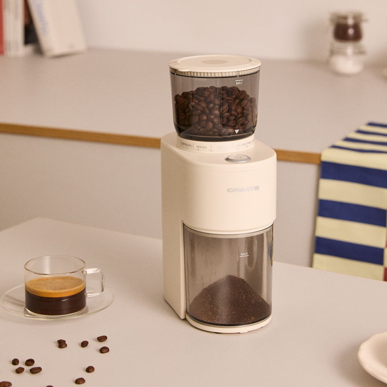 Un molinillo de café blanco con granos arriba y café molido abajo está sobre una mesa junto a una taza de espresso.