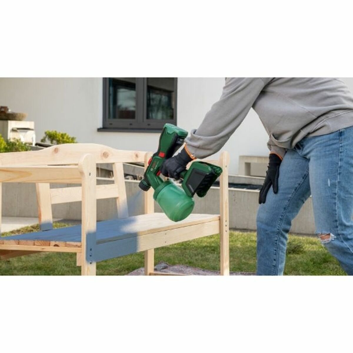 Person malt eine hölzerne Gartenbank mit einem grünen Farbsprühgerät an.