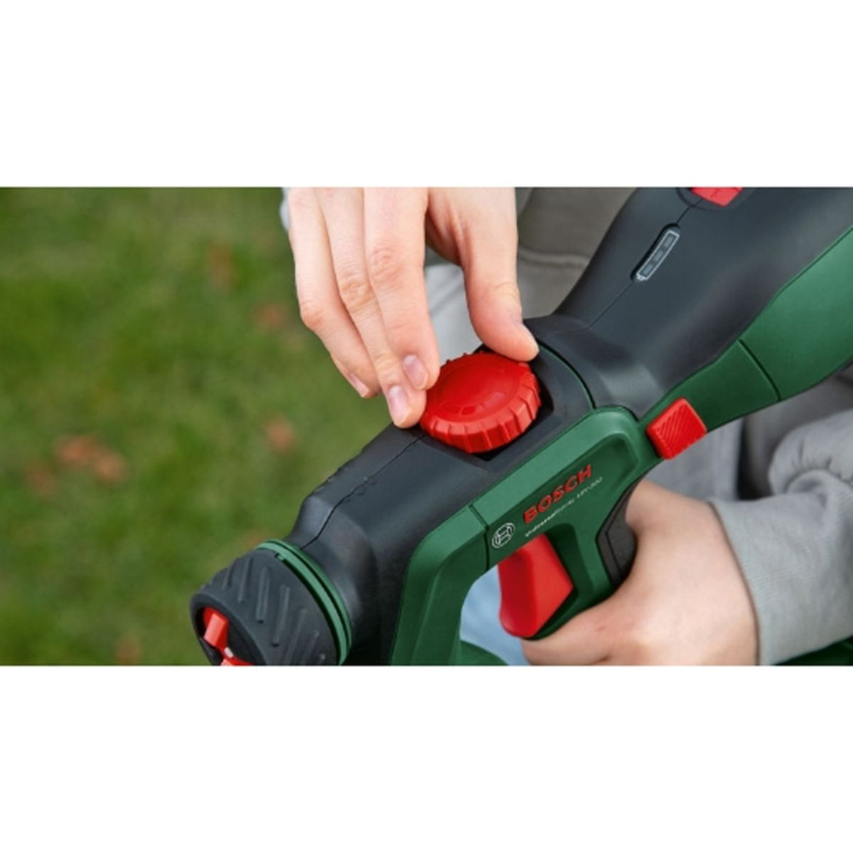 Eine Hand dreht das rote Einstellrad an einem grünen und schwarzen Bosch-Gartengerät vor Gras.