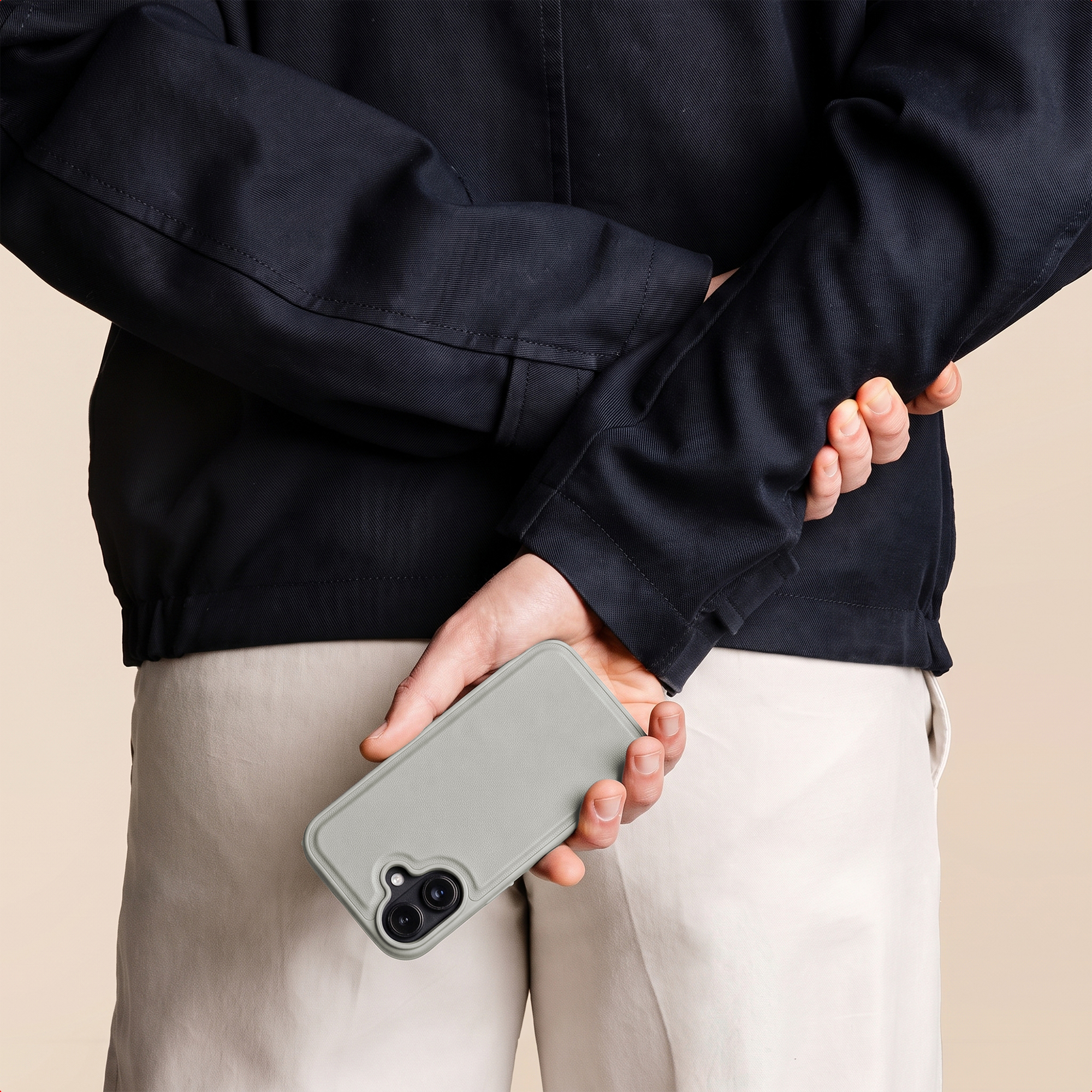 Eine Person in schwarzer Jacke und heller Hose hält ein graues Smartphone hinter ihrem Rücken.