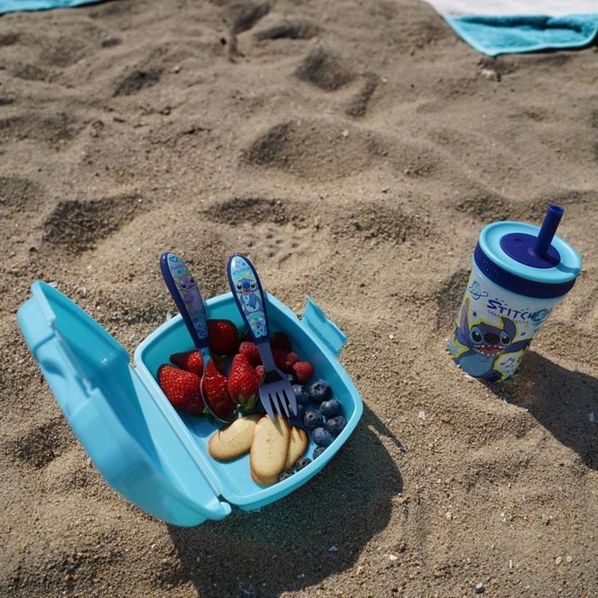 Eine blaue Brotdose mit Obst und Keksen sowie ein Stitch-Becher im Sand.