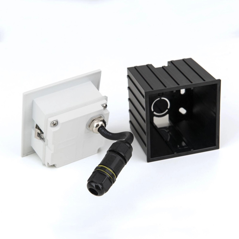Ein weißes Einbauteil mit Kabel und schwarzem Stecker liegt neben einem schwarzen, gerippten Gehäuse auf weißem Grund.