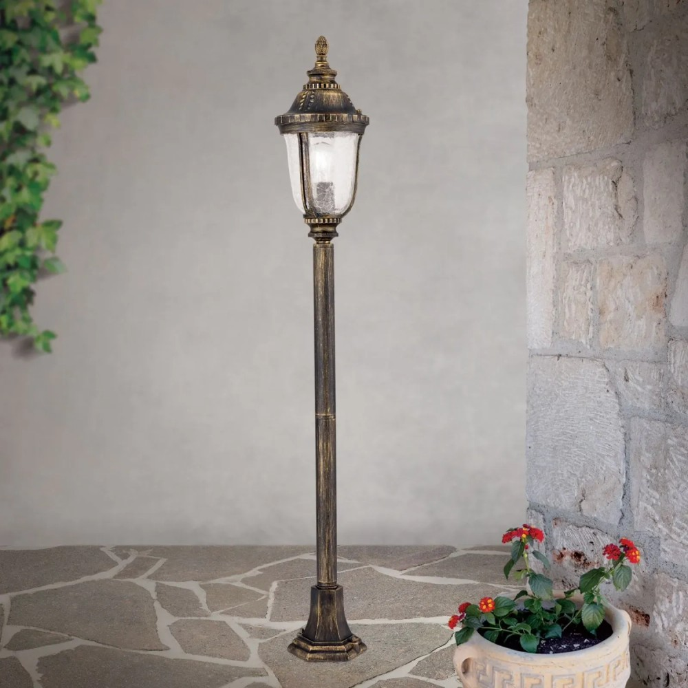 Eine antike Außenstehlampe steht auf Steinpflaster neben einer Steinwand mit Topfpflanze.