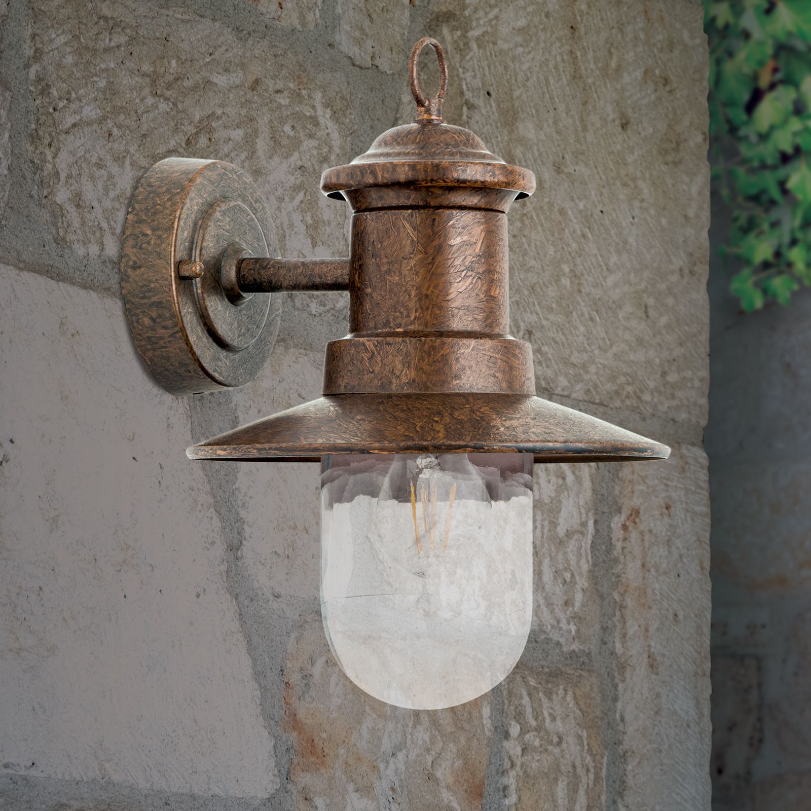 Eine vintage bronzefarbene Wandlampe mit klarem Glas und Glühbirne hängt an einer grauen Steinwand.