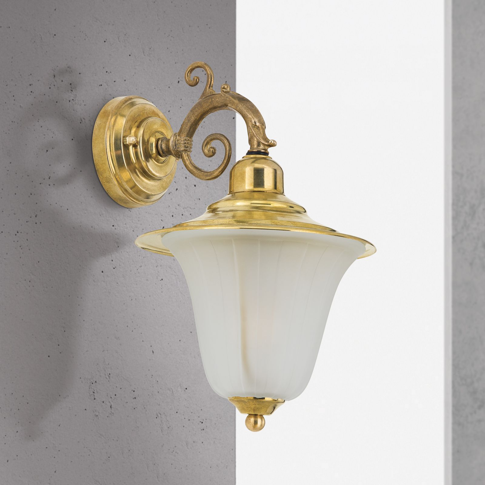 Eine Messing-Wandlampe mit verziertem Schwenkarm und weißem Glasschirm an einer grauen Betonwand.