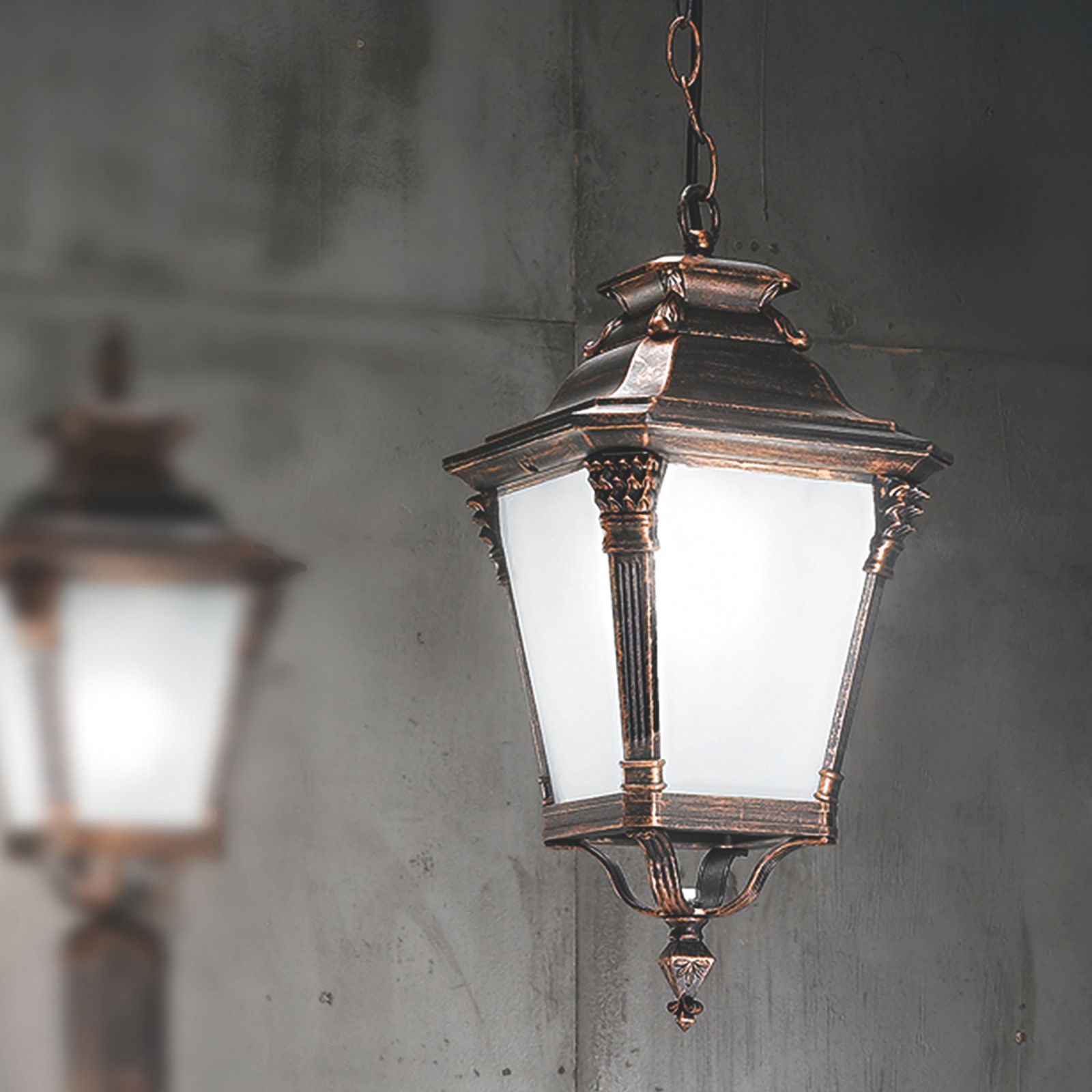 Eine antike, bronzefarbene Hängelampe mit weißem Glas leuchtet vor einer dunklen Betonwand.