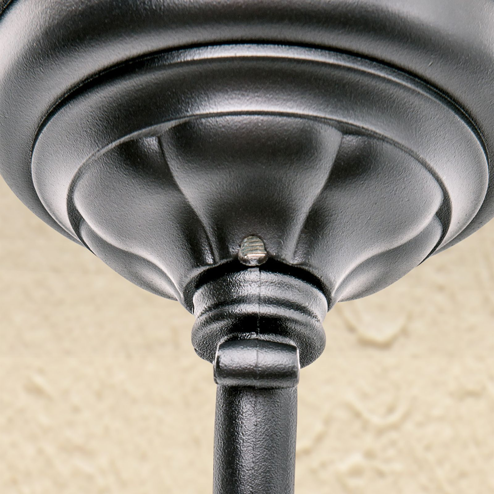Eine schwarze Metall-Leuchtenaufhaengung mit gerilltem Design vor einer strukturierten beigen Wand.