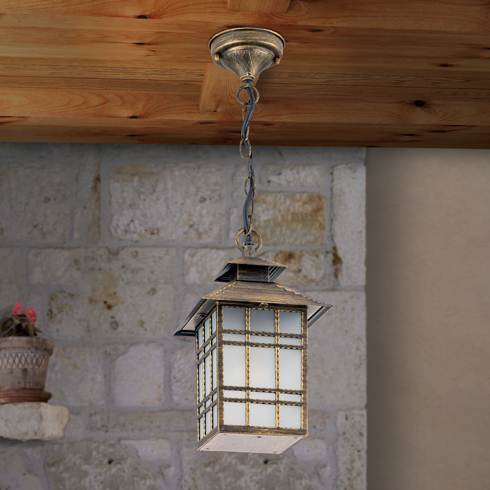 Eine antike, goldfarbene Hängeleuchte aus Metall hängt an einer Kette unter einer Holzdecke vor einer Steinwand.