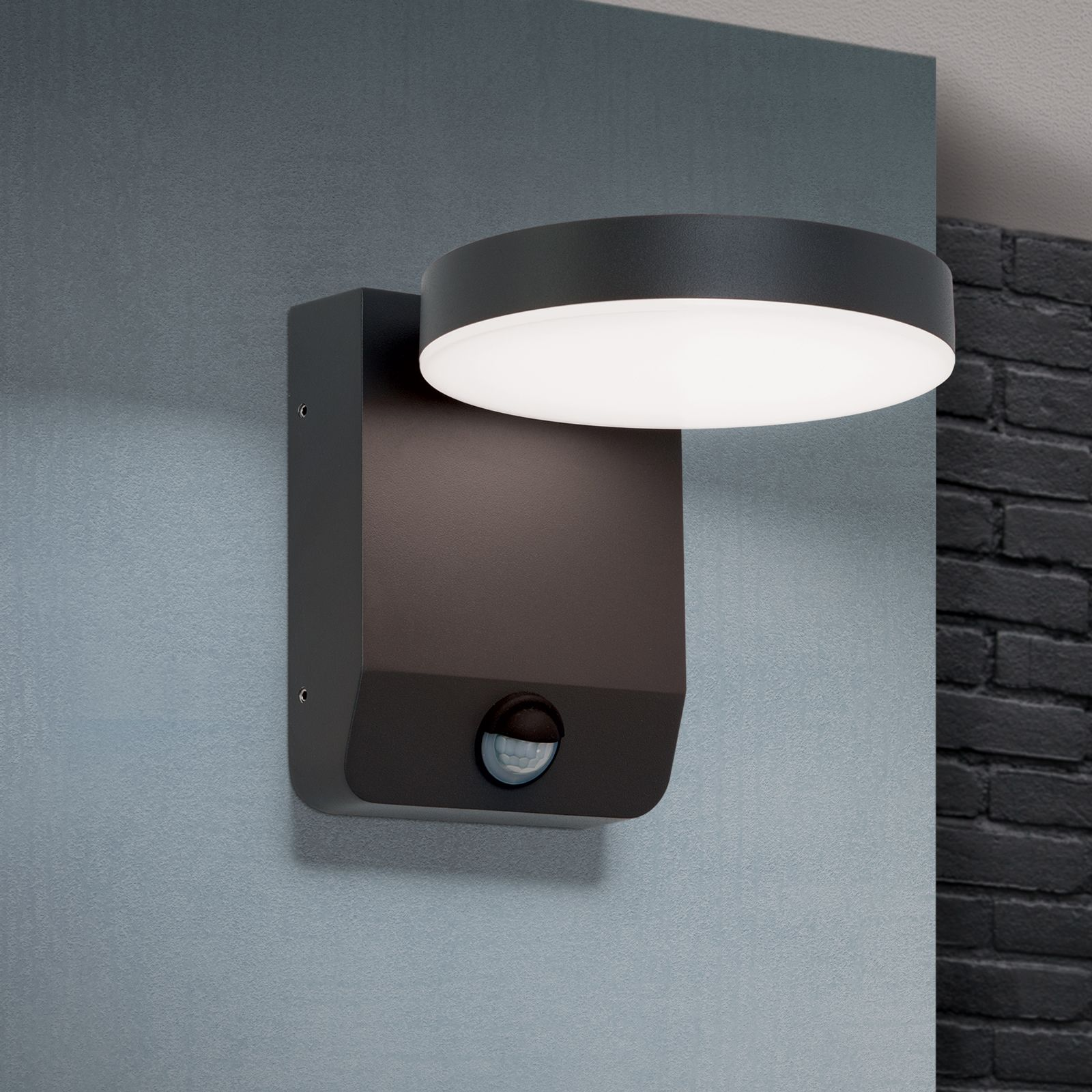 Eine schwarze Wandleuchte mit rundem Leuchtkopf und Bewegungssensor an einer grauen Wand.