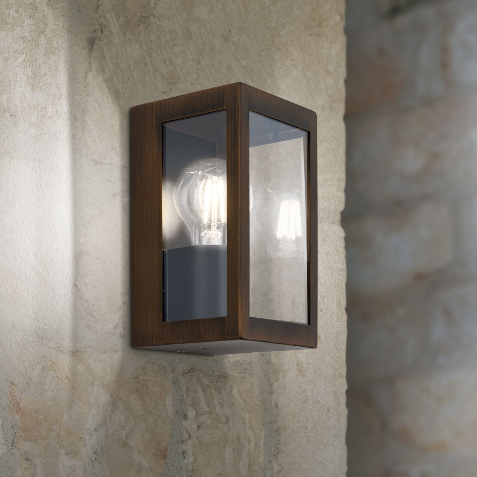 Eine rechteckige braune Metallwandleuchte mit Glasabdeckung und Glühbirne an einer strukturierten Steinwand.
