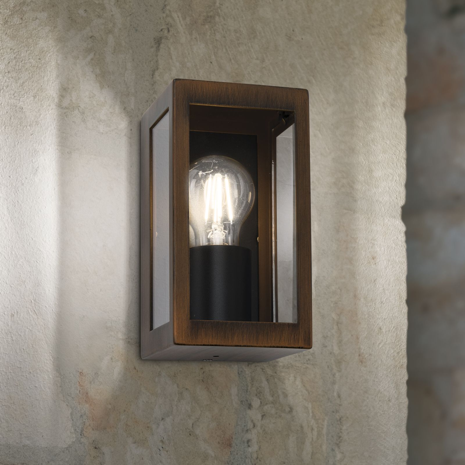 Eine rechteckige Wandleuchte aus Bronze mit Glasseiten und heller Glühbirne an einer rauen Wand.
