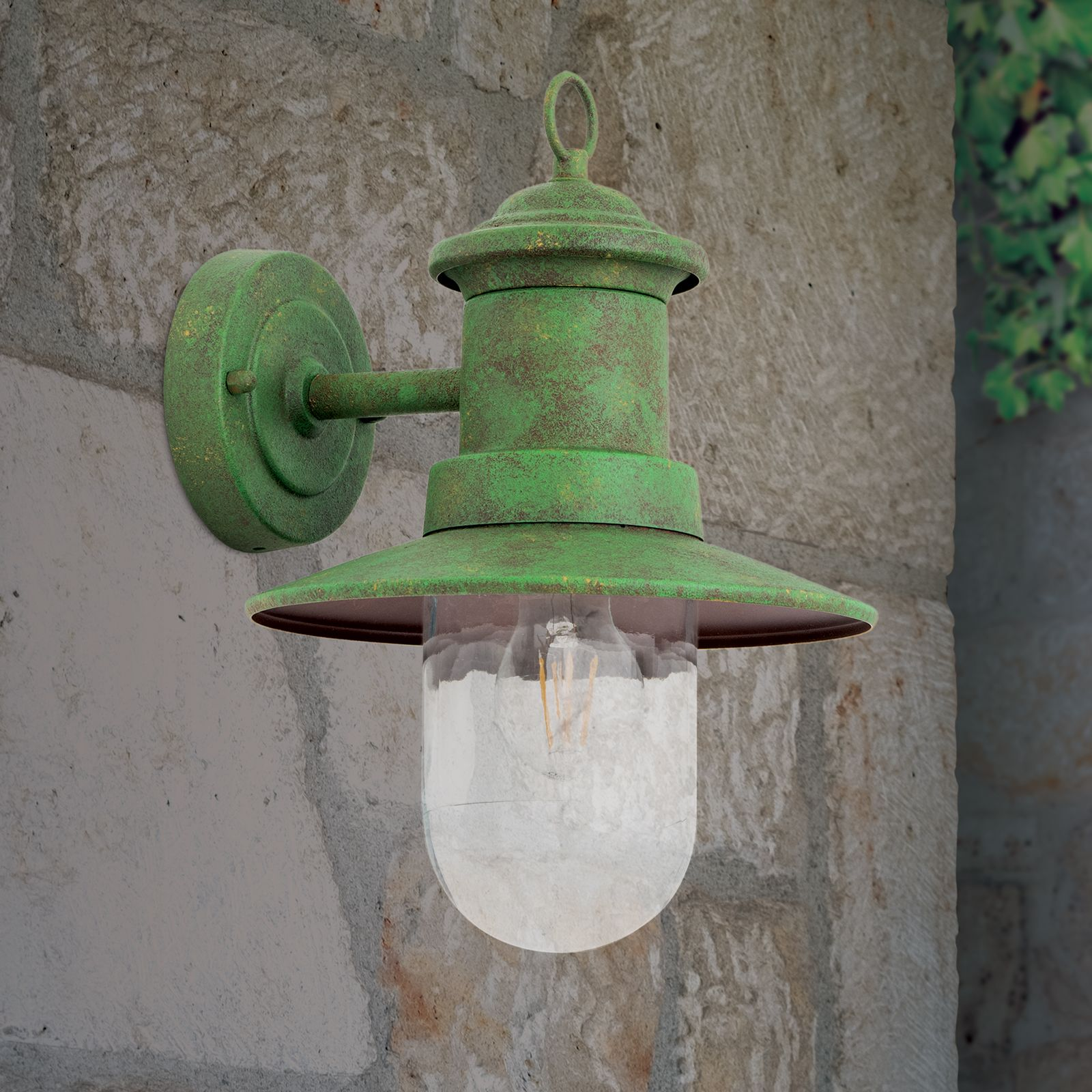 Eine grüne, gealterte Wandlampe mit klarem Glas hst an einer grauen Steinwand befestigt.