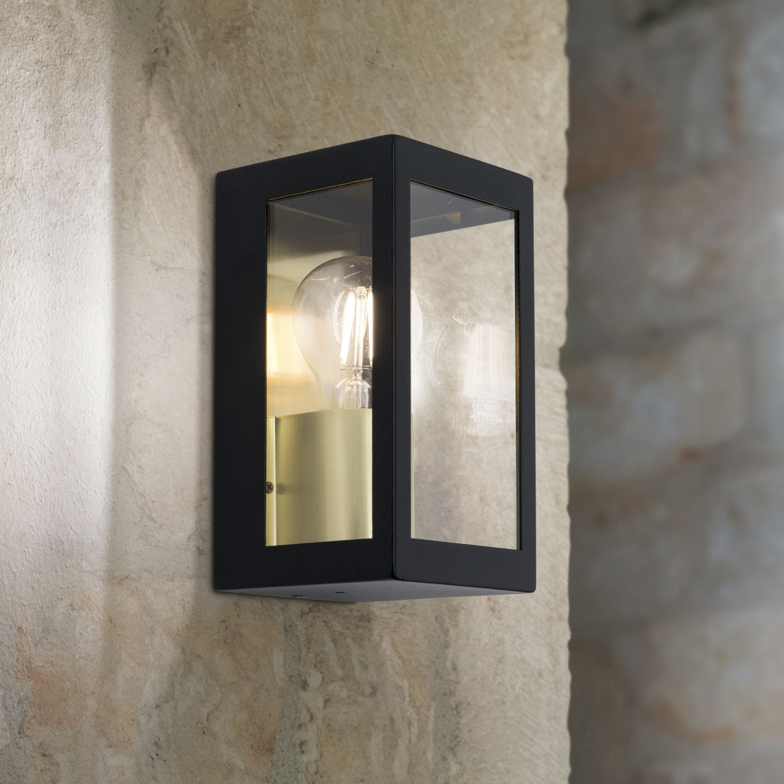 Eine schwarze Wandleuchte mit gläsernem Gehäuse und leuchtender Glühbirne ist an einer Natursteinwand montiert.
