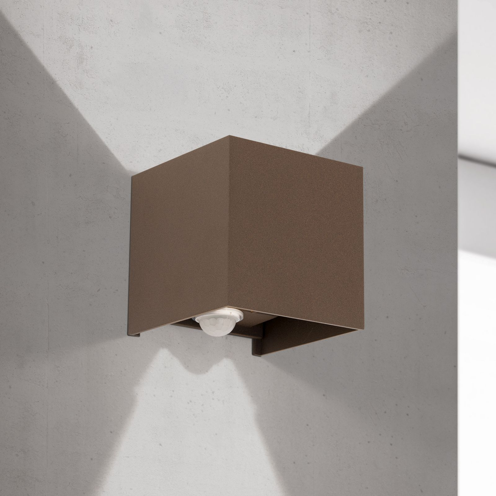 Eine braune, quaderförmige Wandleuchte strahlt Licht nach oben und unten an eine helle Betonwand.