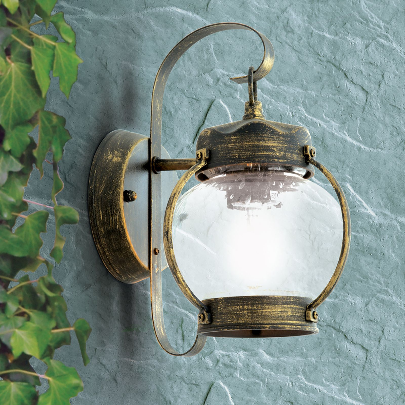 Eine antike gold-schwarze Wandlampe mit runder Glaskugel hängt an einer grauen Wand, links mit Efeublättern.
