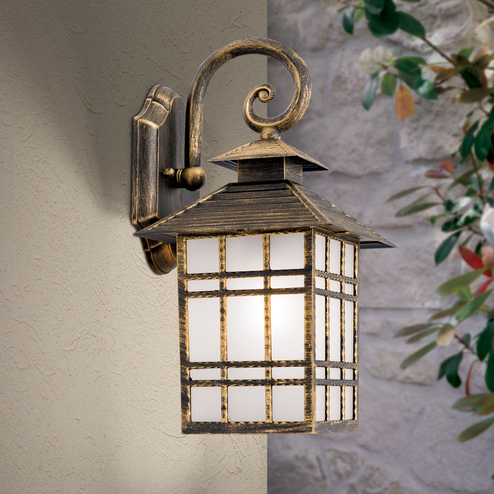 Eine antike schwarze und goldene Wandleuchte mit mattem Glas hängt an einer hellen Wand, im Hintergrund ist Stein und Grün.