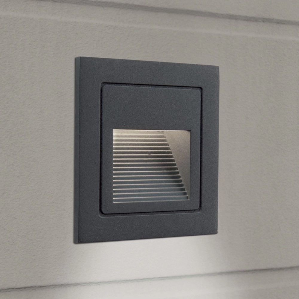 Eine quadratische, dunkle Wandeinbauleuchte mit horizontalen Lamellen wirft sanftes Licht auf eine helle Wand.