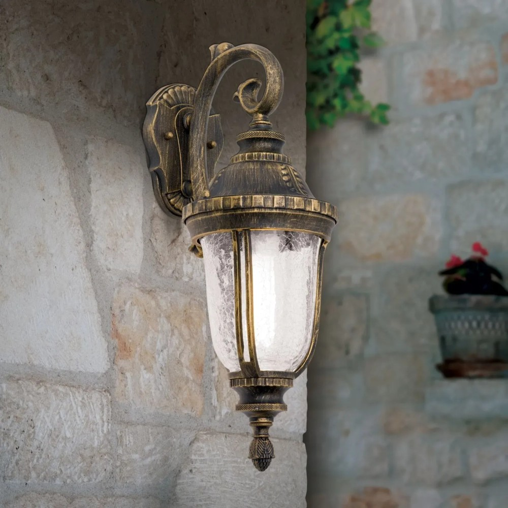 Eine antike goldene Wandlampe mit strukturiertem Glas leuchtet an einer grauen Steinmauer.